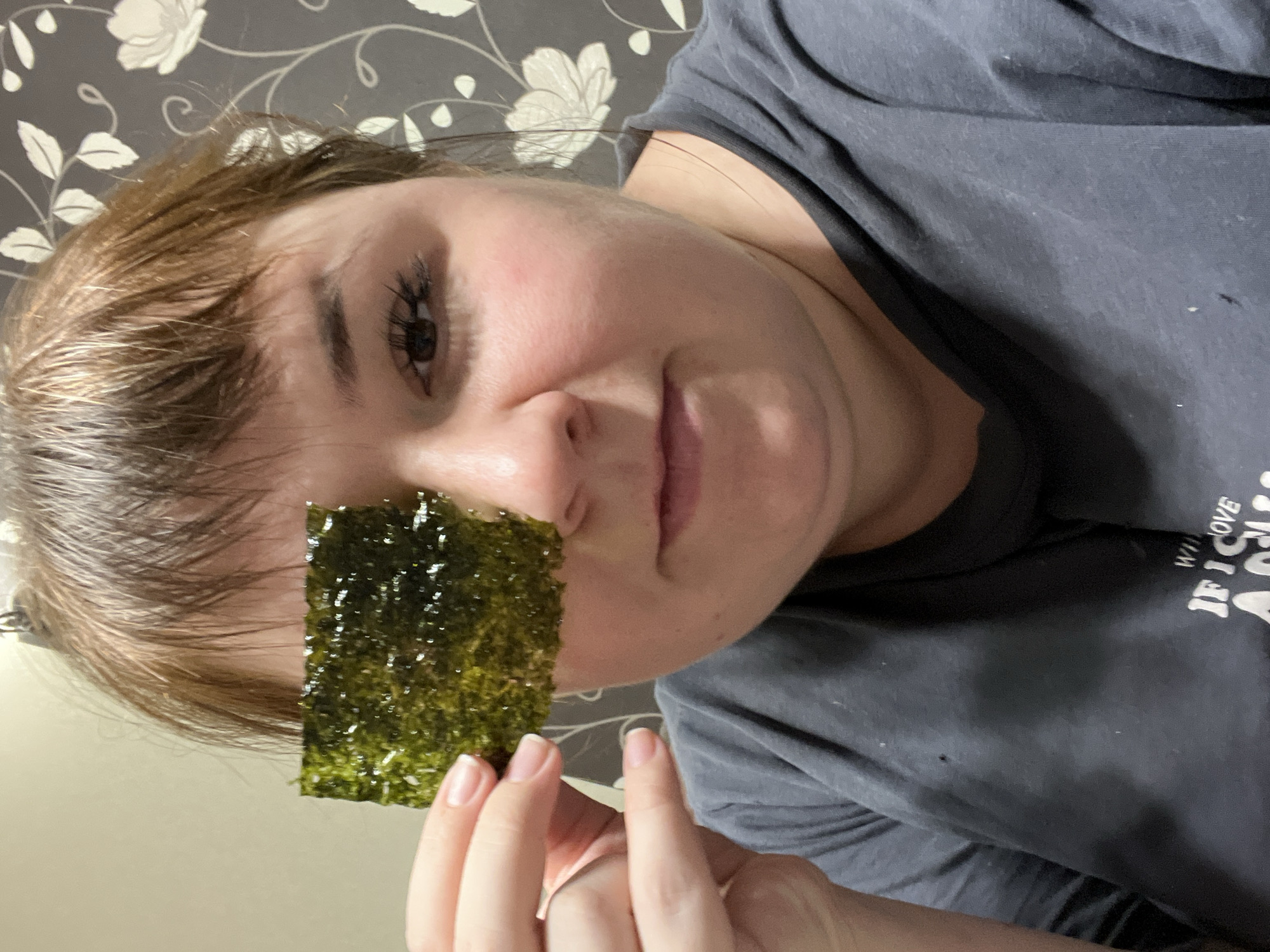 An adult female subject is depicted indoors in Chaikovsky, Russia, shown from the waist up, facing forward. She is holding a rectangular piece of dark green, glossy dried seaweed (nori) over her left eye with her left hand. Her right eye, featuring a dark iris and extended eyelashes, is visible and directed towards the viewer. The subject has light brown hair with bangs and maintains a neutral expression with closed lips. She is wearing a dark grey t-shirt, which displays a partial white inscription on the lower right side and has several small white specks and a dark mark. A simple ring is present on her left ring finger. The background features a wall covered with dark grey wallpaper adorned with a repeating pattern of white flowers and leaves.