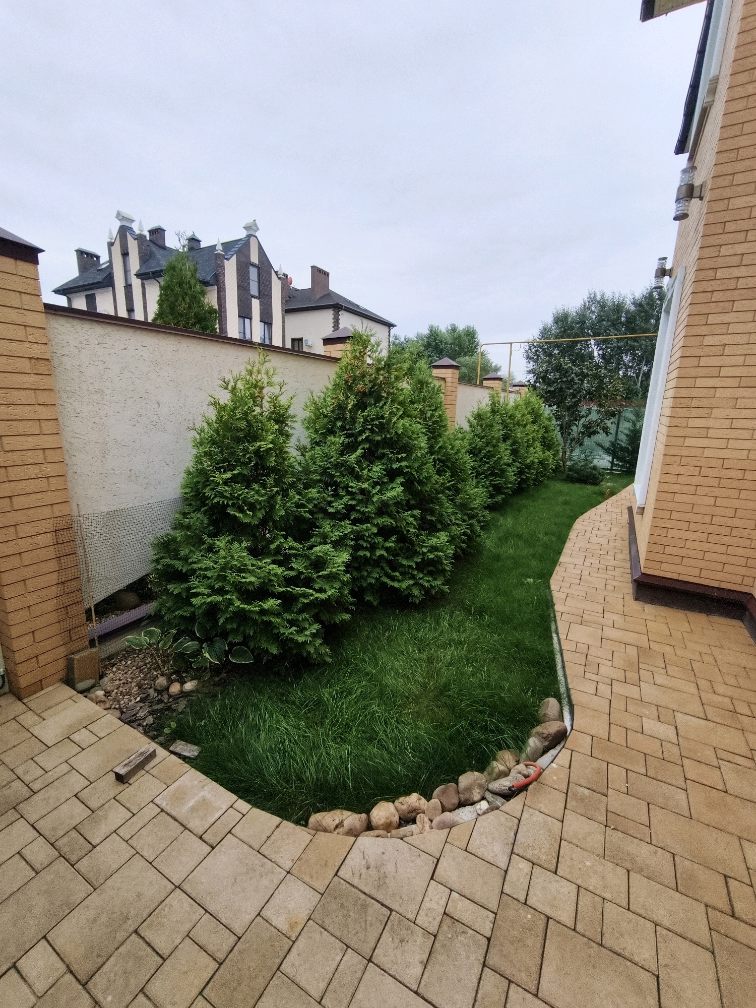 A view of a backyard with a paved walkway and a well-maintained lawn bordered by a row of evergreen trees. In the background, a light-colored textured wall separates the yard from a row of distinctive houses with dark roofs and white trim. The houses have a modern, somewhat ornate architectural style. The sky is overcast. A portion of a brick house is visible on the right side of the image. The scene appears to be a residential area, possibly in Krasnodar, Russia, given the architectural style of the houses.