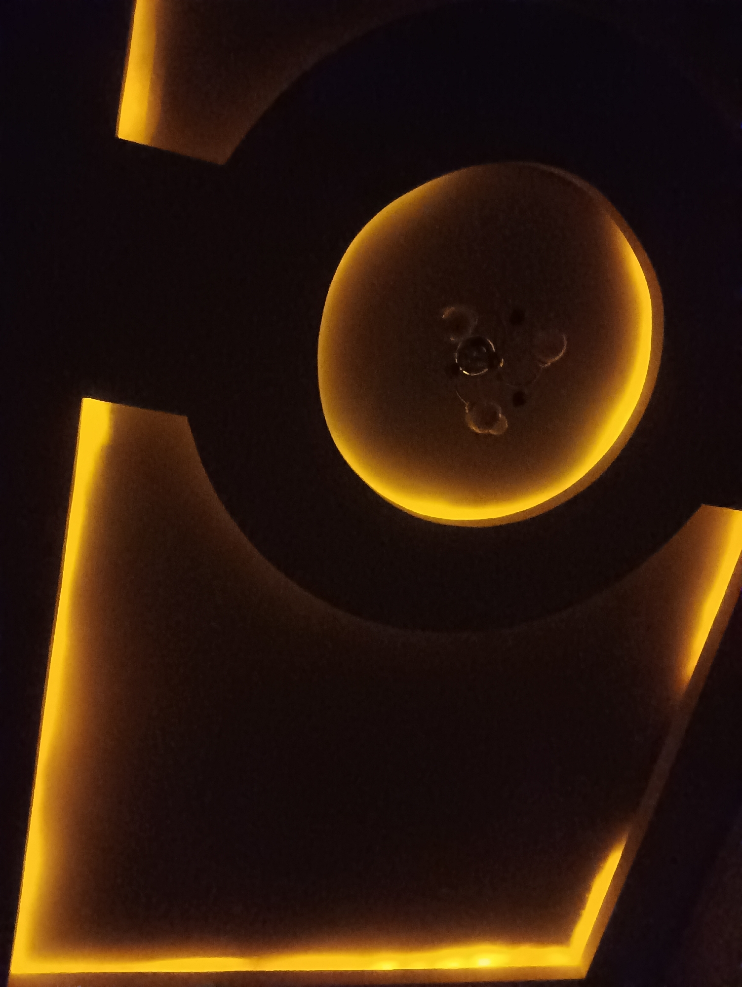 The image displays a modern interior design element, likely a ceiling or wall feature, characterized by dark surfaces illuminated by integrated yellow LED lighting. The design incorporates geometric shapes, including a prominent curved recess and angular projections. Within the curved recess, a light fixture with multiple bulbs is visible, though it is partially obscured by the surrounding architecture and the lighting conditions. The overall aesthetic suggests a contemporary and possibly sophisticated space, with the warm glow of the lighting creating a dramatic effect. No specific geographical markers or elements are visible to indicate the city of Angren, Uzbekistan.