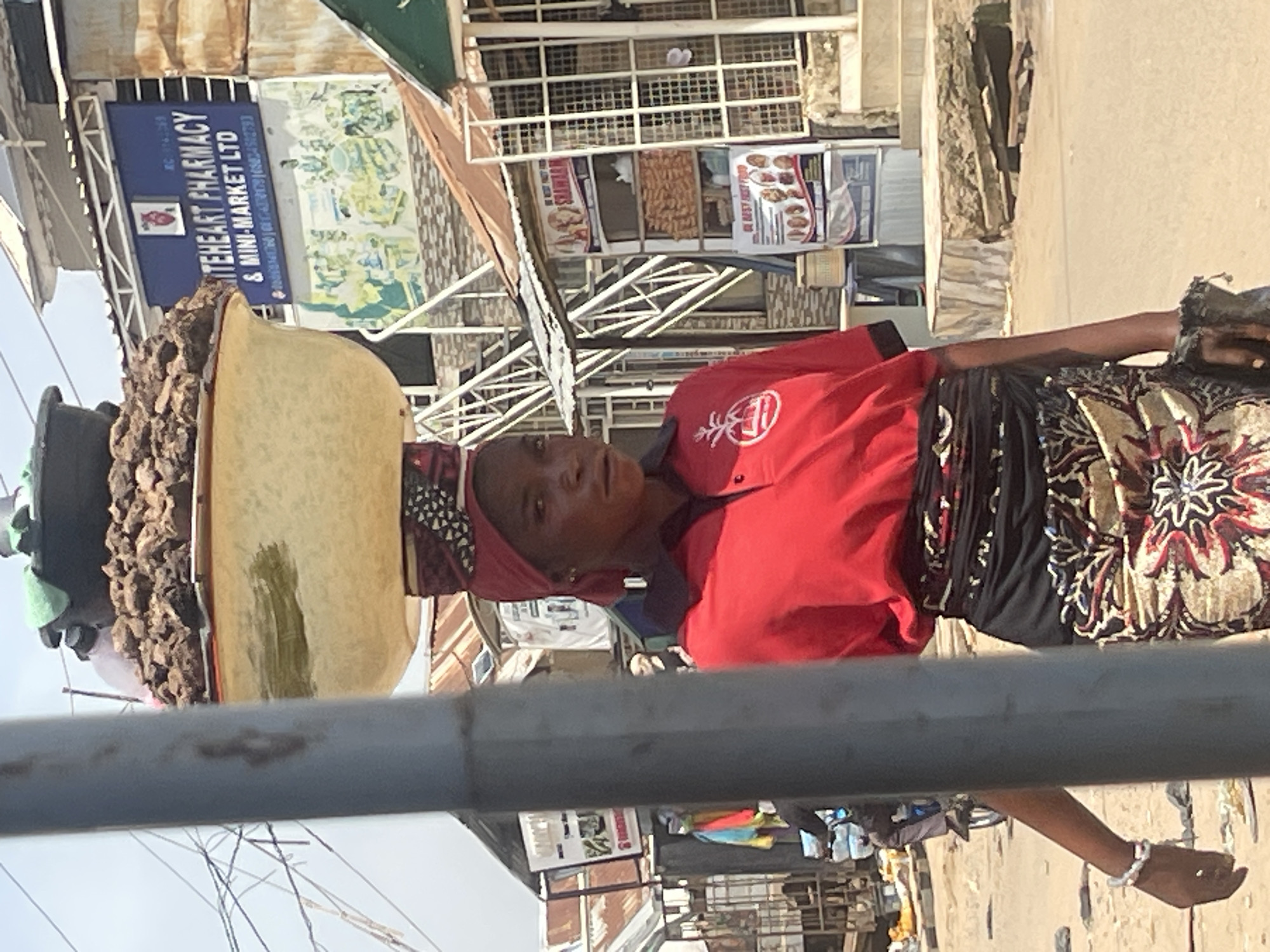 The image shows a woman in a busy outdoor market, likely in Abuja, Nigeria. She is carrying a large, round basket of goods on her head. The basket appears to be filled with dried food items, possibly nuts or spices, and is balanced on a cushion or cloth wrapped around her head. She is wearing a bright red polo shirt with a white logo and a colorful, patterned skirt. Her head is covered with a pink or red headscarf.
The background reveals a bustling marketplace with various stalls and shops. A prominent blue sign reads "WHITEHEART PHARMACY & MINI-MARKET LTD". Other signs and displays suggest retail businesses selling food items and other goods. The overall scene depicts a typical day in a vibrant African market, with the woman actively participating in commerce, likely as a vendor or transporter of goods. The lighting suggests it is daytime, and the scene is outdoors, possibly on a street or open-air market area. The woman's expression is somewhat neutral, perhaps focused on her task or observing her surroundings. The presence of other people and the general activity imply a lively and active environment.