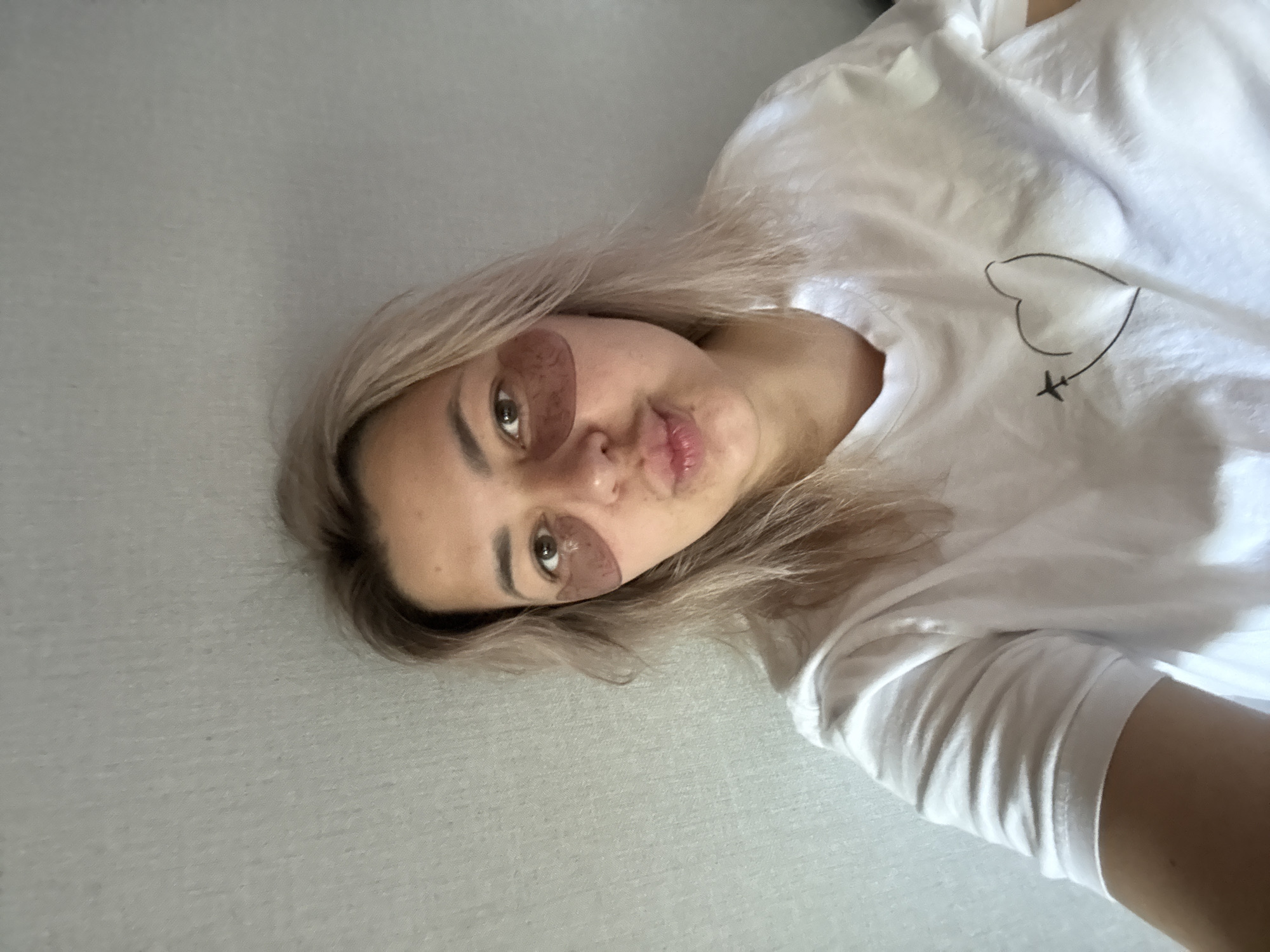 A woman with light blonde hair and light brown eyes is taking a selfie. She is wearing under-eye patches and a white t-shirt with a heart and airplane design. The background appears to be a textured wall. The image does not provide any information about the location in Birobidzhan, Russia.