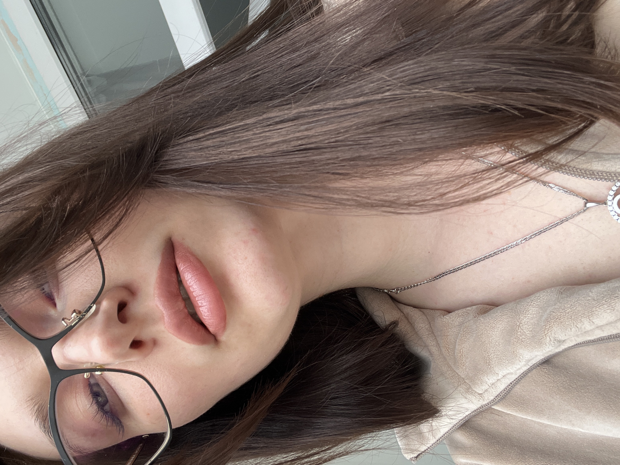 A close-up portrait shows a young woman, likely indoors in Tyumen, Russia, with the image rotated 90 degrees clockwise. Her long, dark brown hair cascades across a significant portion of the frame. She wears black-framed eyeglasses, revealing one eye with what appears to be dark purple or blue mascara. Her full lips, adorned with pinkish-nude lipstick, are slightly parted. A silver-colored chain necklace with a circular pendant is visible around her neck. She is dressed in a light beige or cream-colored jacket with a textured, possibly suede or fleece-like, surface. The background is softly blurred, suggesting an indoor setting with a window, indicated by muted grey and white tones. The lighting appears natural, suggesting daytime. Her expression is neutral, possibly contemplative, as she poses for the camera. No visible text is present.