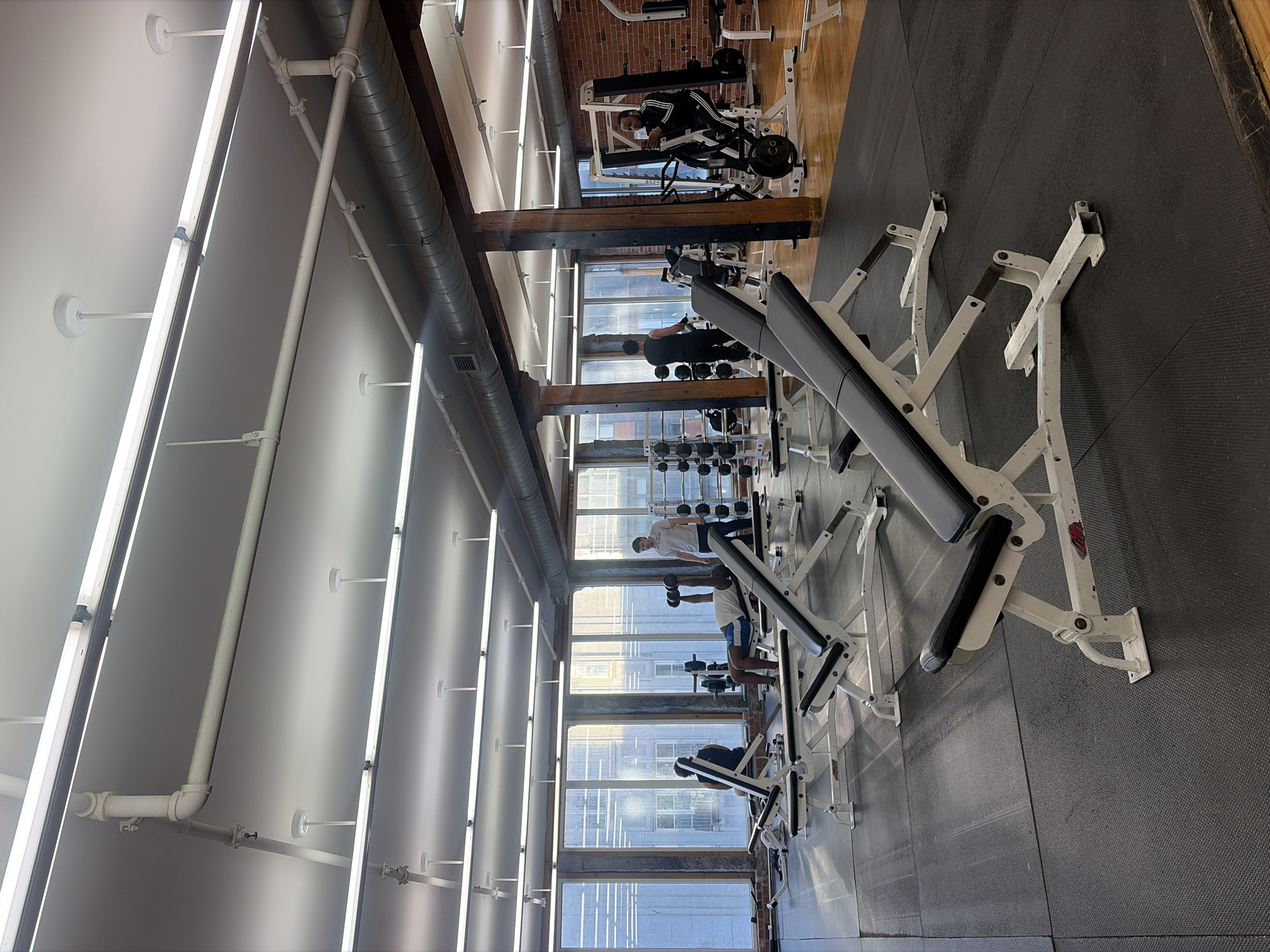 The image depicts the interior of a gym, presumably located in a city, given the urban landscape visible through the large windows. There is no information in the picture to suggest that this is in Montreal, Canada, or anywhere else specifically. The gym features rows of weight benches, each appearing to be in use by individuals engaged in weightlifting exercises. The gym space is illuminated by long, fluorescent lights affixed to the ceiling, and the floor is covered with black rubber mats, commonly used in gyms for shock absorption and to protect the underlying surface. The structure of the building is partly exposed, revealing wooden beams and a gray ventilation system. There's also a portion of a brick wall visible in the background.