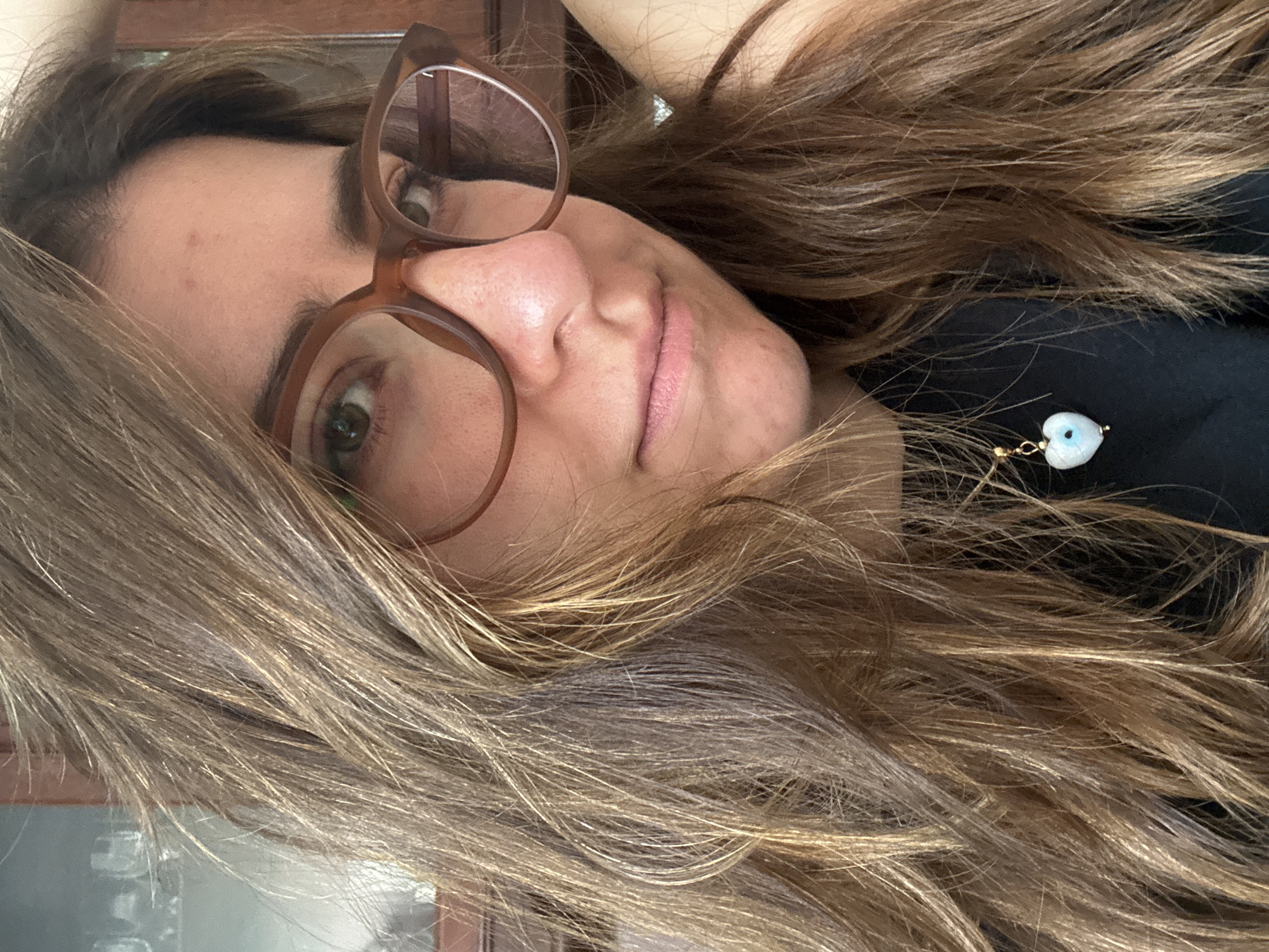 A woman with brown hair and brown-rimmed glasses is lying down, facing the camera. She is wearing a black top with a small heart-shaped charm on a delicate gold chain. The background is slightly blurred, and part of a wooden frame is visible on the left. The location is indeterminable from this image.