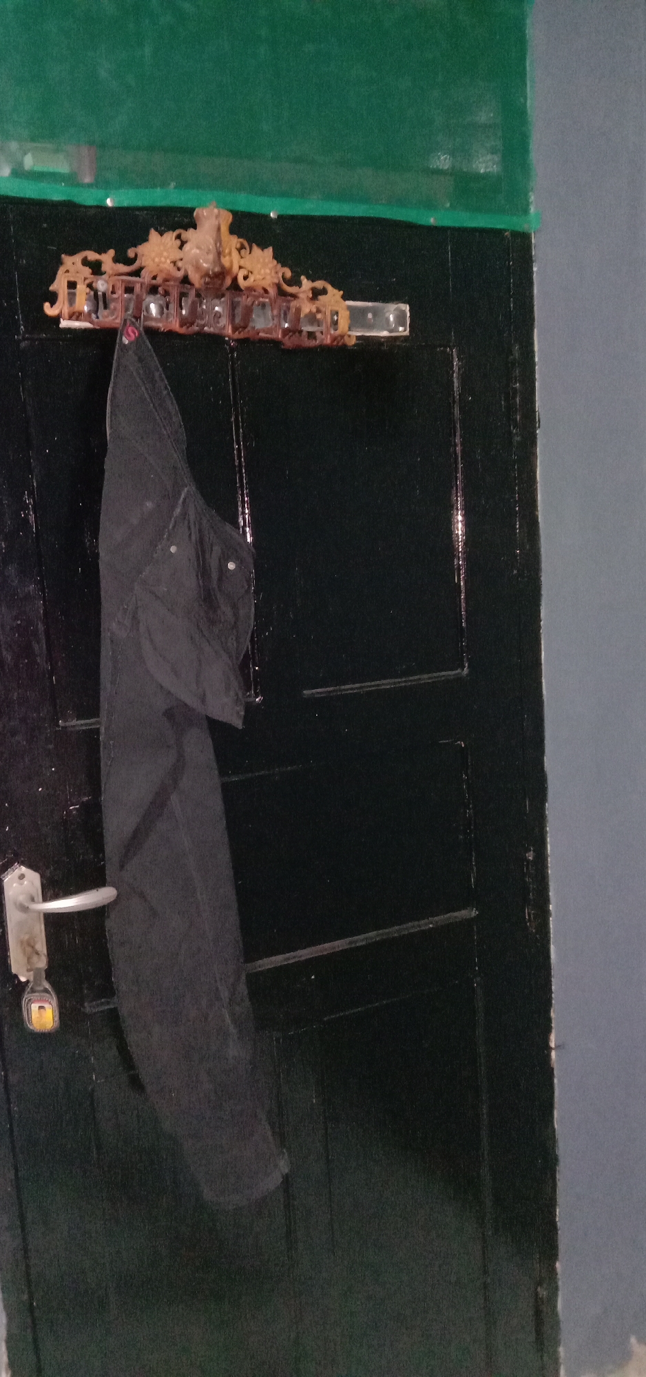 The image depicts a close-up of a dark green painted door with a silver door handle and a small yellow tag. Hanging from a decorative, ornate wooden hanger attached to the top of the door is a pair of black trousers. The hanger features intricate carvings, including what appears to be a face or figure at its center, and some rectangular elements that could be decorative inlays or part of its structure. The wall to the right of the door is a muted gray color, and the area above the door is a solid dark green. The scene appears to be indoors, in a residential or utilitarian space. There is no indication of the city of Kendari, Indonesia, within the visual information provided in the image.