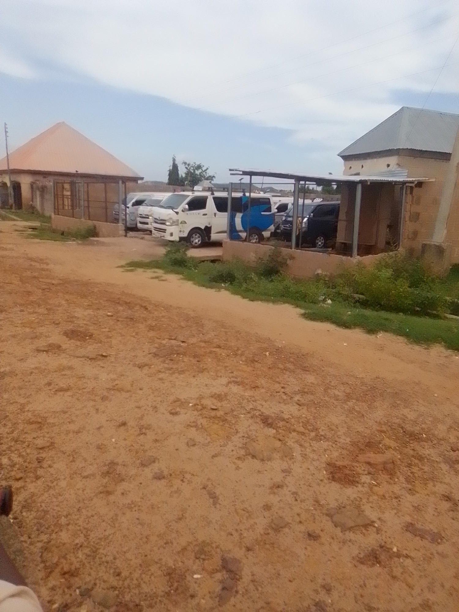 In Wuro Galadima, Nigeria, a dirt lot adjacent to buildings is occupied by several parked vehicles. A light-colored, multi-paneled structure with a peaked roof is situated on the left. In the center, multiple white vans are parked under a covered shelter. To the right, a brown brick building with a gray metal roof is visible. The ground is unpaved and composed of reddish-brown dirt with scattered small stones and patches of green vegetation. The sky is overcast with scattered clouds. No people are visible in the image.