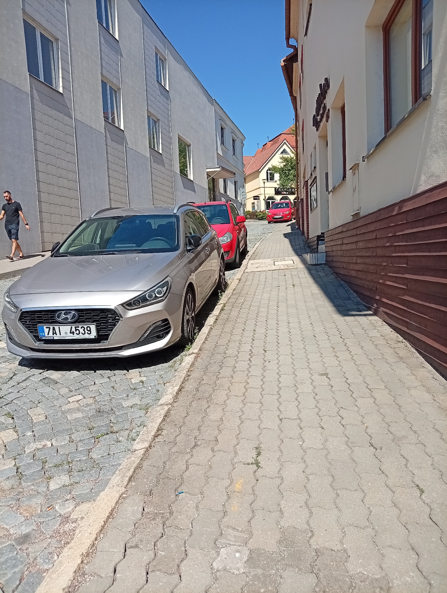 The image depicts a narrow street in Tišnov, Czechia, with buildings lining both sides. On the left, a modern, grey building with multiple windows dominates the foreground. A man in a black t-shirt and dark shorts is walking on the cobblestone pavement in front of it. A silver Hyundai station wagon is parked on the cobblestones, its front facing the camera. A red hatchback is parked behind the Hyundai. The right side of the street features a building with a white facade and brown trim, alongside a narrow sidewalk paved with interlocking concrete blocks. Further down the street, red cars and more traditional buildings with tiled roofs are visible under a clear blue sky. The scene suggests a sunny day in a European town.