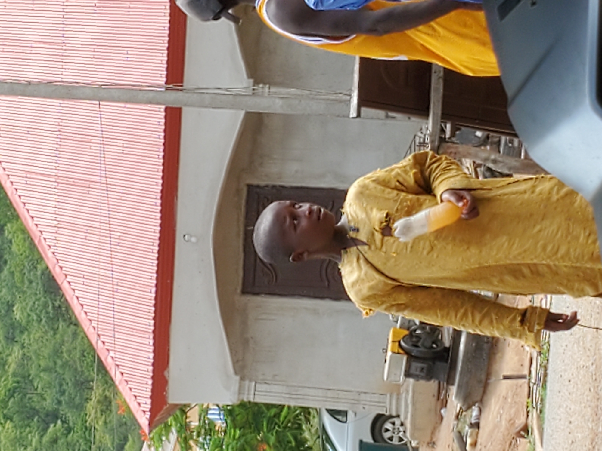 In Mararaba, Nigeria, an individual wearing a mustard-yellow garment stands outdoors. The person holds a small, translucent yellow object in their left hand. The background features a building with a red roof, a grey door, and white walls. A partial view of a white vehicle and some greenery is also visible.