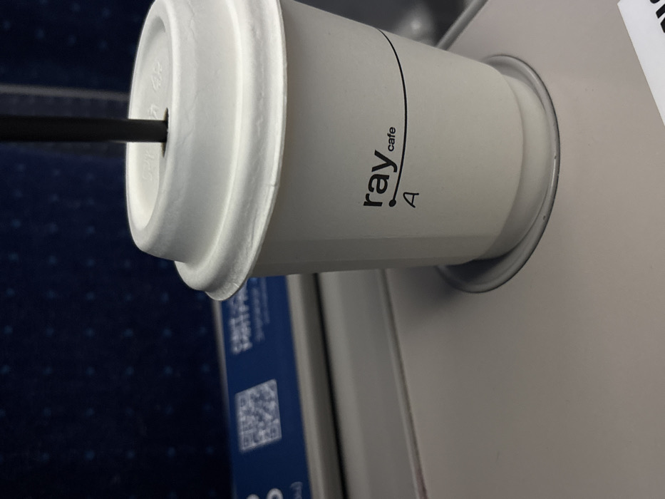 A white paper cup, branded "ray cafe A," rests in a silver cup holder. The cup has a white lid with a black straw inserted. Part of a dark blue textured surface is visible in the background, along with a partially visible blue and white sign featuring a QR code. The cup holder is attached to a light gray, possibly plastic, surface.