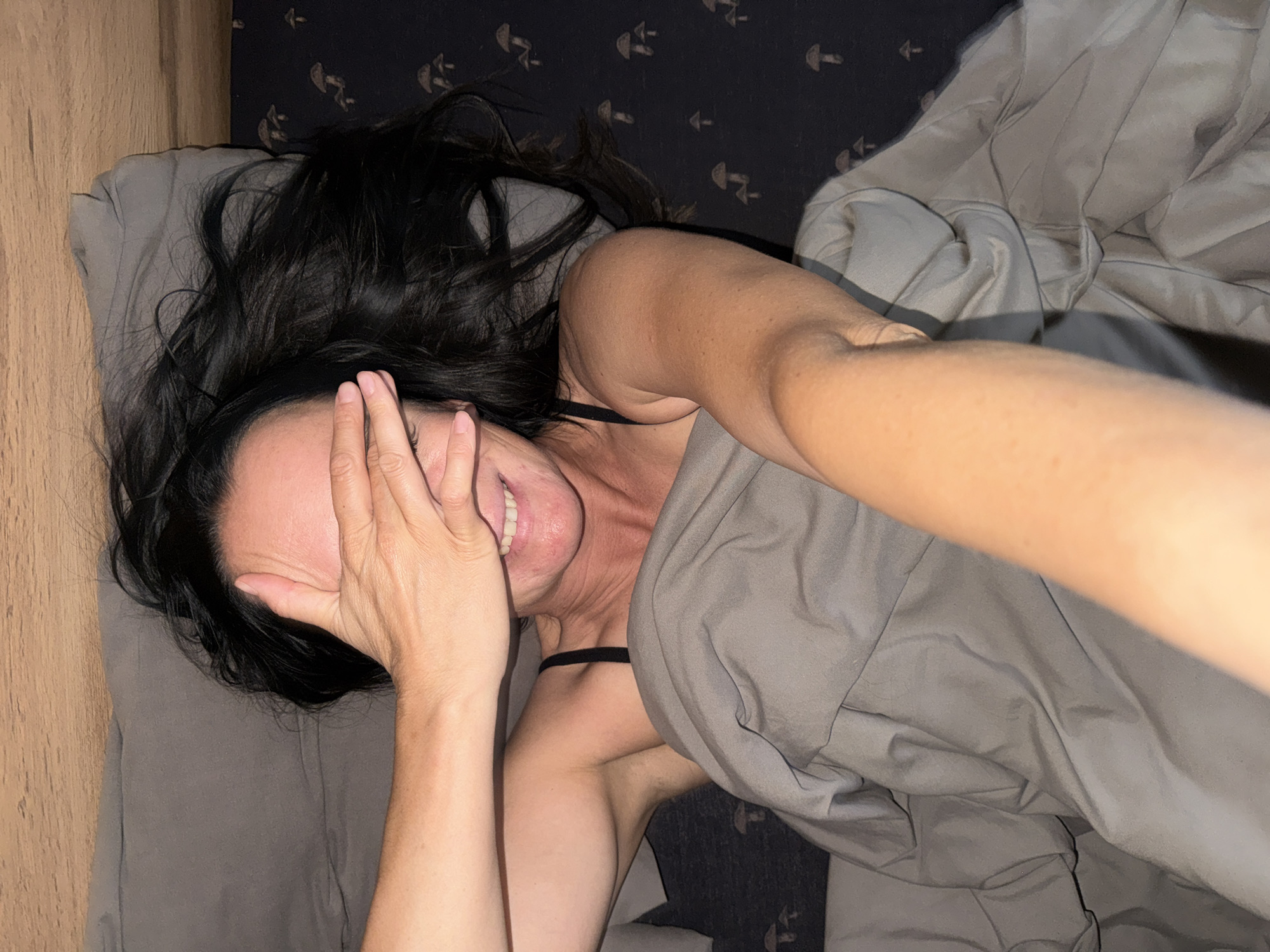 A woman with long, dark hair lies in bed, covering her eyes with her right hand and smiling. She is partially covered by a gray comforter. The background shows a dark patterned sheet with mushroom designs and a wooden headboard. The scene appears to be in a bedroom. This image was taken in Chaikovsky, Russia.