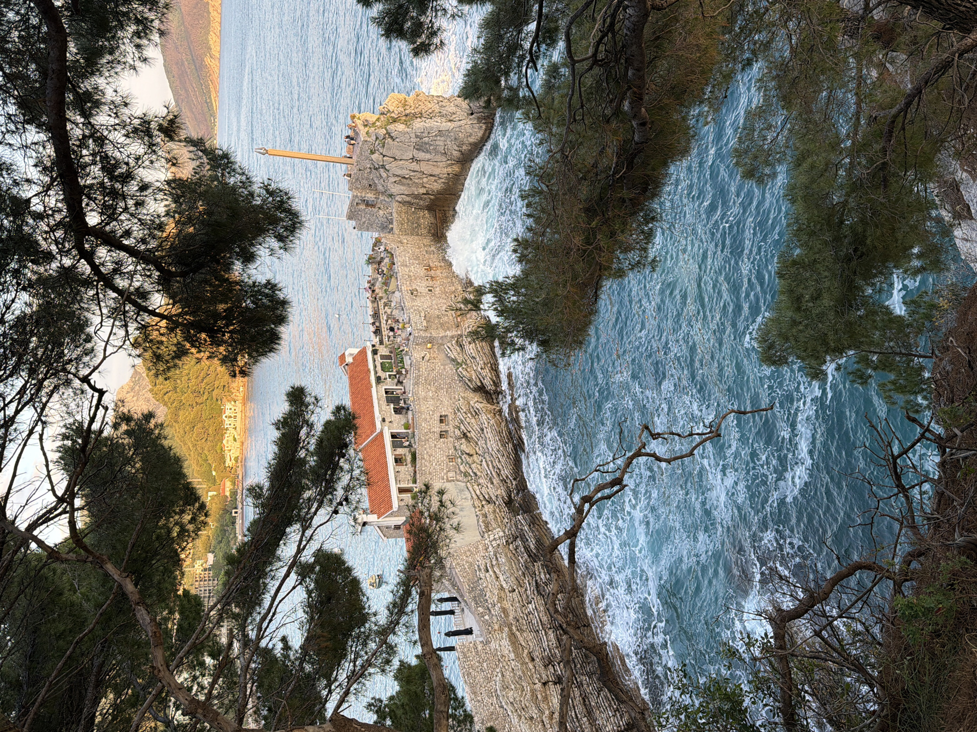 A top-down view shows a coastal scene in Petrovac centar, Montenegro. The horizon features a mountain ridge, and below lies the sea, characterized by turbulent blue waters. A rocky outcrop with a structure, possibly a building with a red-tiled roof and walls, juts into the water. Adjacent to the building is a stone terrace with seating. Trees frame the view from both sides. A tall, wooden structure stands at the edge of the rocky outcrop. The image is rotated 90 degrees.