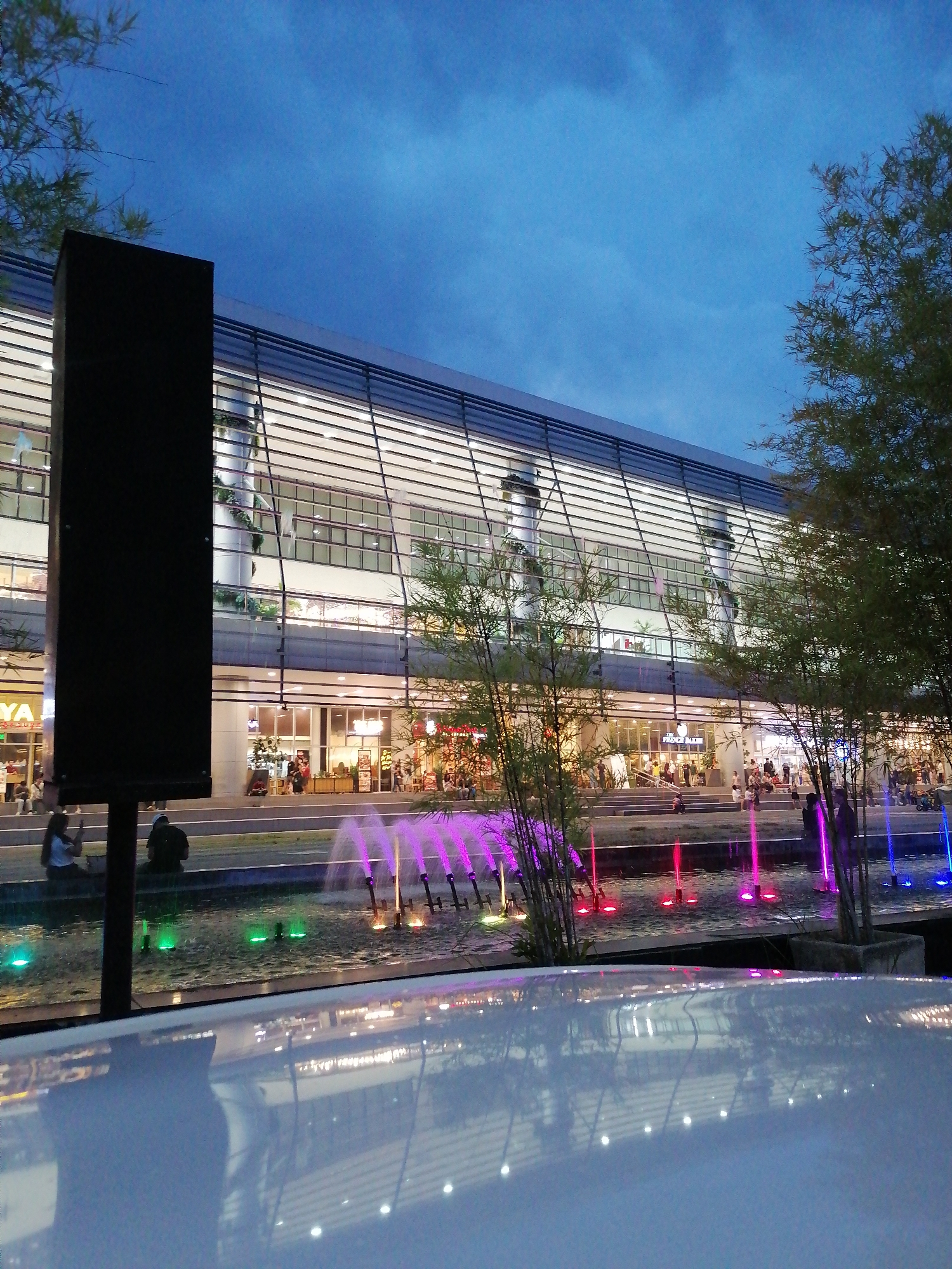 The image captures a nighttime view of a modern building in Davao City, Philippines, likely a commercial establishment given the presence of shops and people. The building features a sleek, horizontal design with extensive glass windows that are illuminated from within. In front of the building, a water feature with colored lights—primarily purple, pink, and blue—is active, with water jets creating a dynamic visual effect. Several people are visible, some seated near the water feature and others walking or standing in front of the building's entrance, suggesting a public space. Bamboo-like plants are strategically placed, adding a touch of nature to the urban landscape. In the foreground, the reflective surface of a white object, possibly a vehicle or a piece of furniture, occupies the lower portion of the frame, mirroring the lights and the building's facade. The sky is a deep blue, indicative of dusk or early night. The overall scene conveys a lively and modern atmosphere.