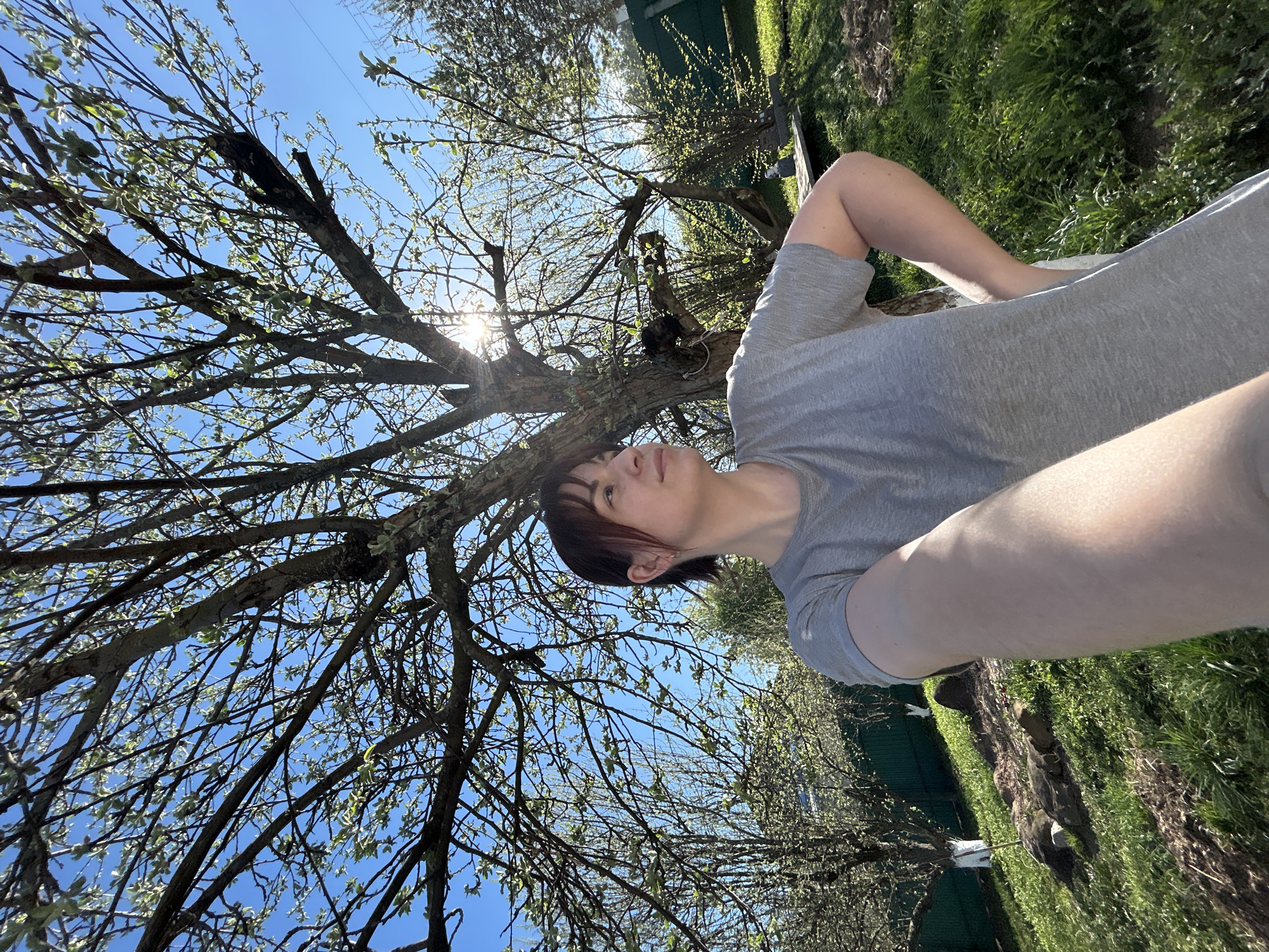 A person with short, dark hair and a grey t-shirt is in the lower right of the frame. The person appears to be taking a selfie. Behind the person is a tree with bare branches against a bright blue sky and sun. Green grass and a partial view of a green structure are visible. The scene appears to be outdoors. The location is Фоки, Russia.