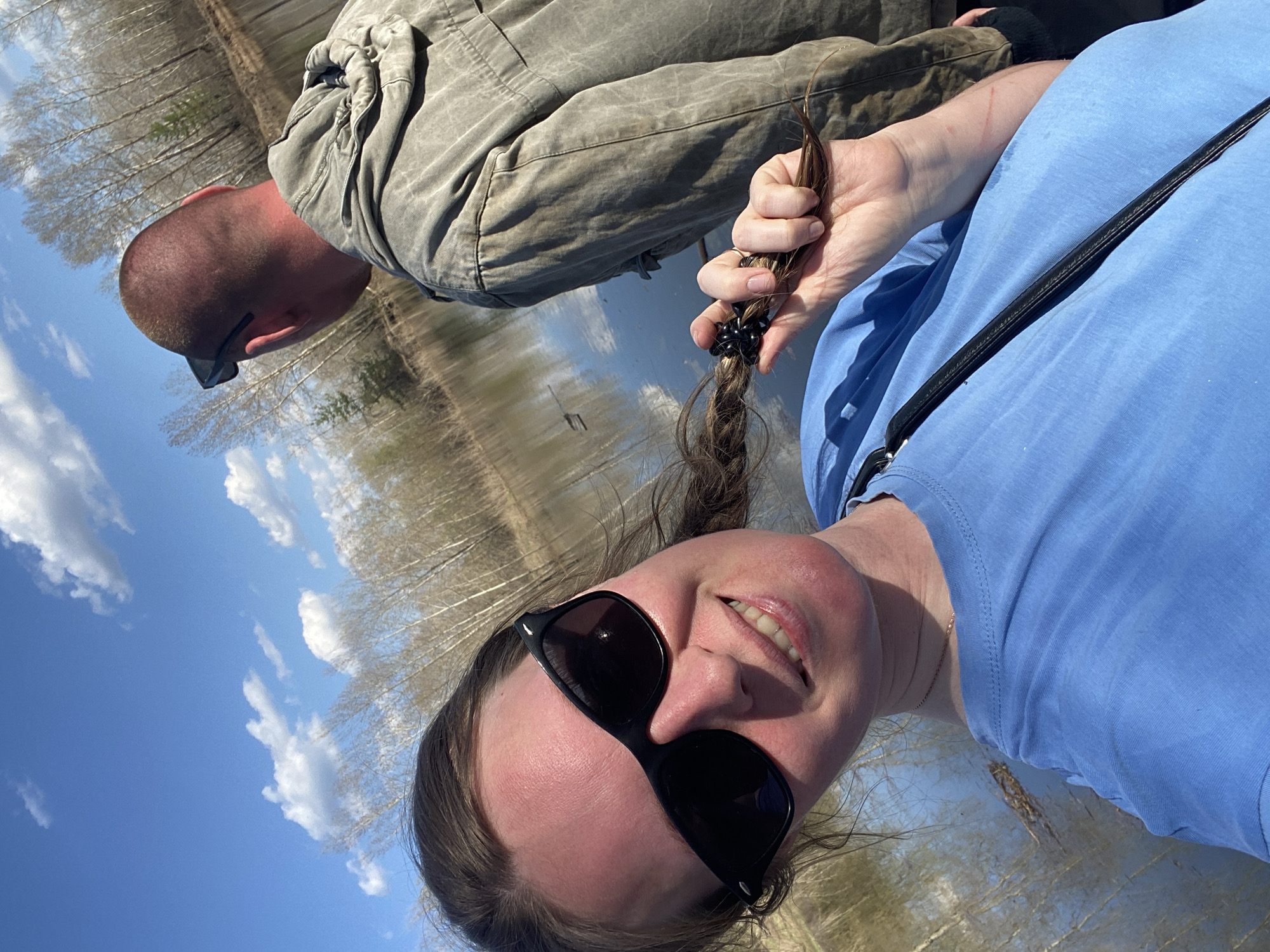The image depicts a selfie taken by a woman wearing sunglasses and a light blue t-shirt. She is holding a section of her braided hair in her hand, which is tied with black elastics. A man wearing sunglasses and a light brown jumpsuit is visible in the background. The background also shows a body of water reflecting the sky and a line of trees on the distant shore. The location is possibly in the city of Chaikovsky, Russia, near a body of water reflecting a partly cloudy sky.