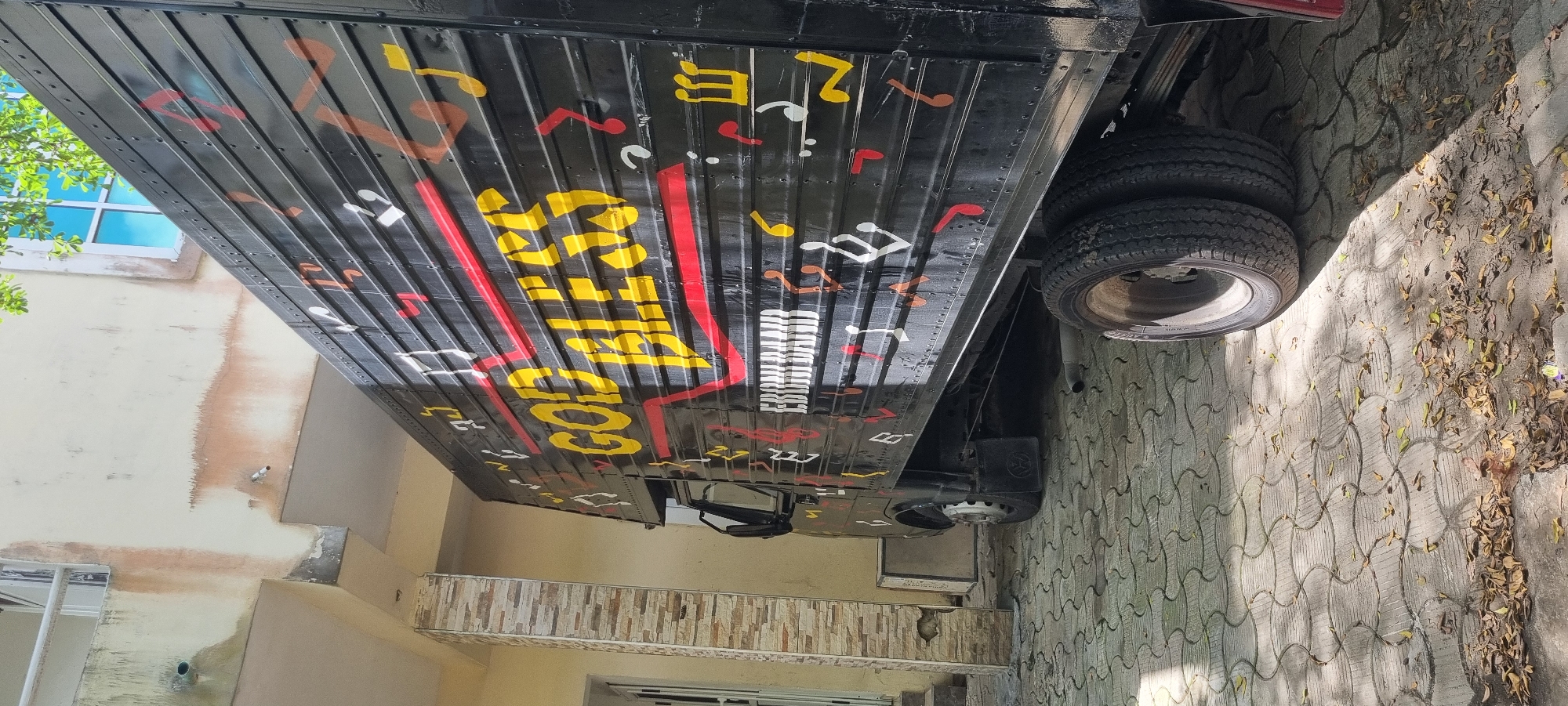 The image depicts a black truck parked in what appears to be a residential area in Lekki, Nigeria. The truck's cargo area is adorned with colorful graffiti-style art, including musical notes and the word "GOLDEN." The setting includes a building with light-colored walls and a window visible in the upper left corner. The ground is paved with interlocking blocks, and scattered leaves suggest an outdoor environment. The lighting indicates it is daytime, with shadows cast by the truck and surrounding structures. No people are visible in the shot.
