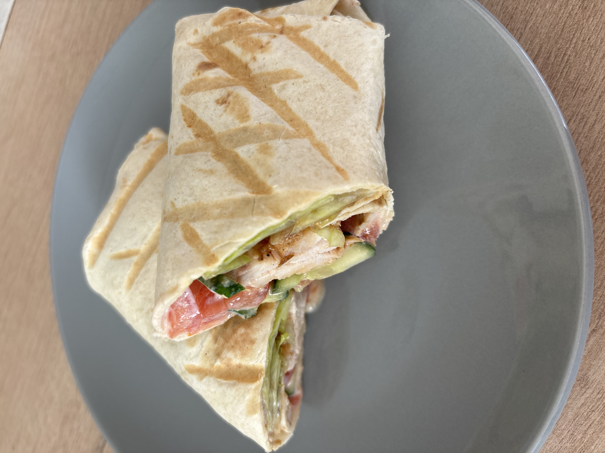 The image shows two chicken wraps on a gray plate, which is resting on a light brown surface. The wraps appear to contain chicken, tomato, cucumber, and a white sauce. The tortillas are marked with a light brown pattern. The photograph does not offer sufficient information to confirm the location as Chaikovsky, Russia.