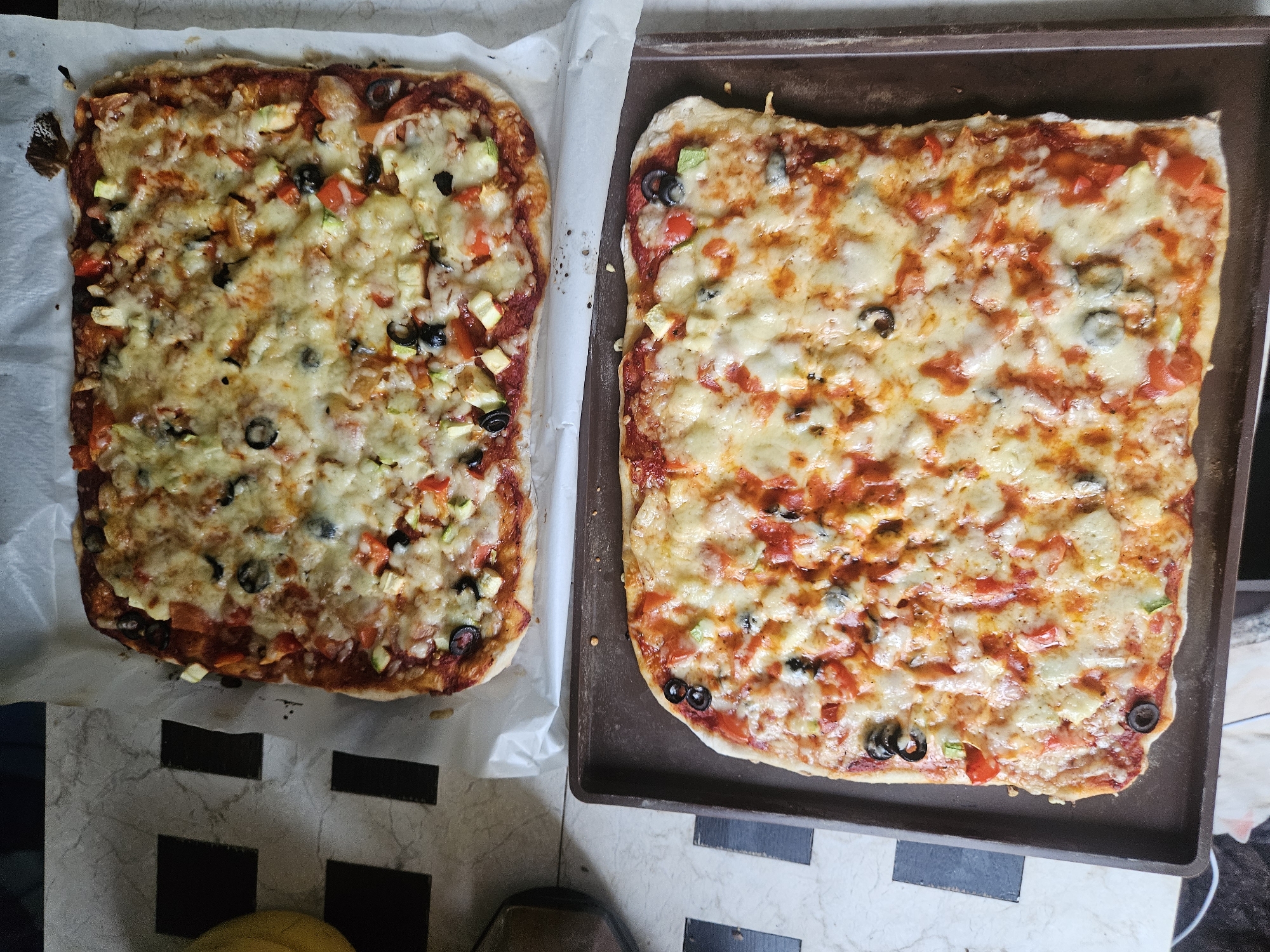 Two square homemade pizzas are displayed on a tabletop. The pizza on the left rests on white parchment paper, while the pizza on the right is on a dark brown baking sheet. Both pizzas are topped with tomato sauce, melted cheese, and scattered vegetable toppings, including black olives, diced red peppers, and zucchini. The tabletop surface is partially visible with a black and white checkered pattern. The location of the scene is not identifiable as Rodniki, Russia, from the visual information provided.