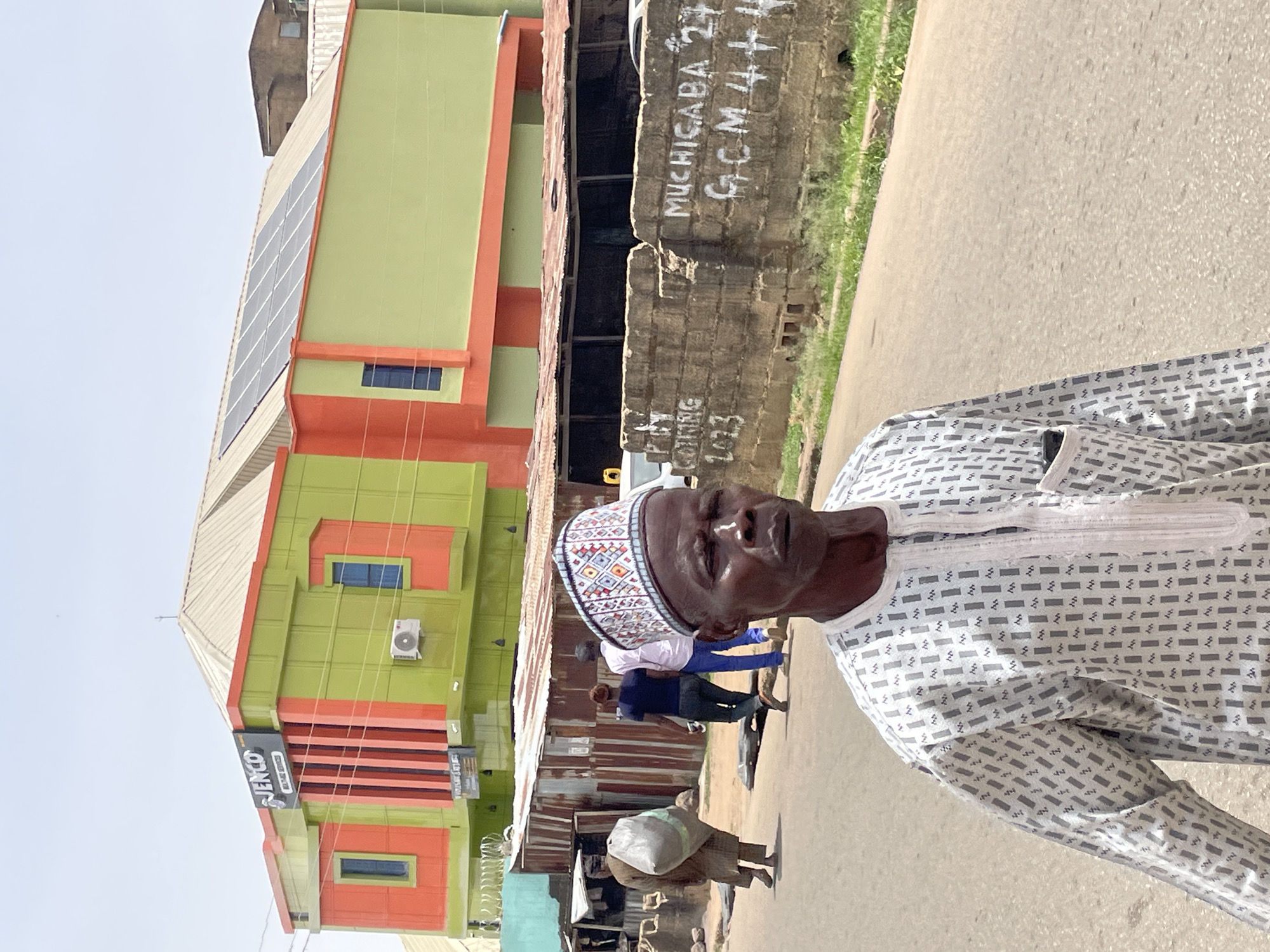 The image shows a man in a patterned white and gray traditional outfit and a decorated cap. He is standing on a street in Jos, Nigeria. Behind him is a building painted green and orange with "JENCO" written on a sign. Solar panels are visible on the upper portion of the building. Other individuals are walking in the background. A brick wall with "MUCHICABA 27" and "GCM 4 +1 +" written on it stands to the right. The street appears to be paved.