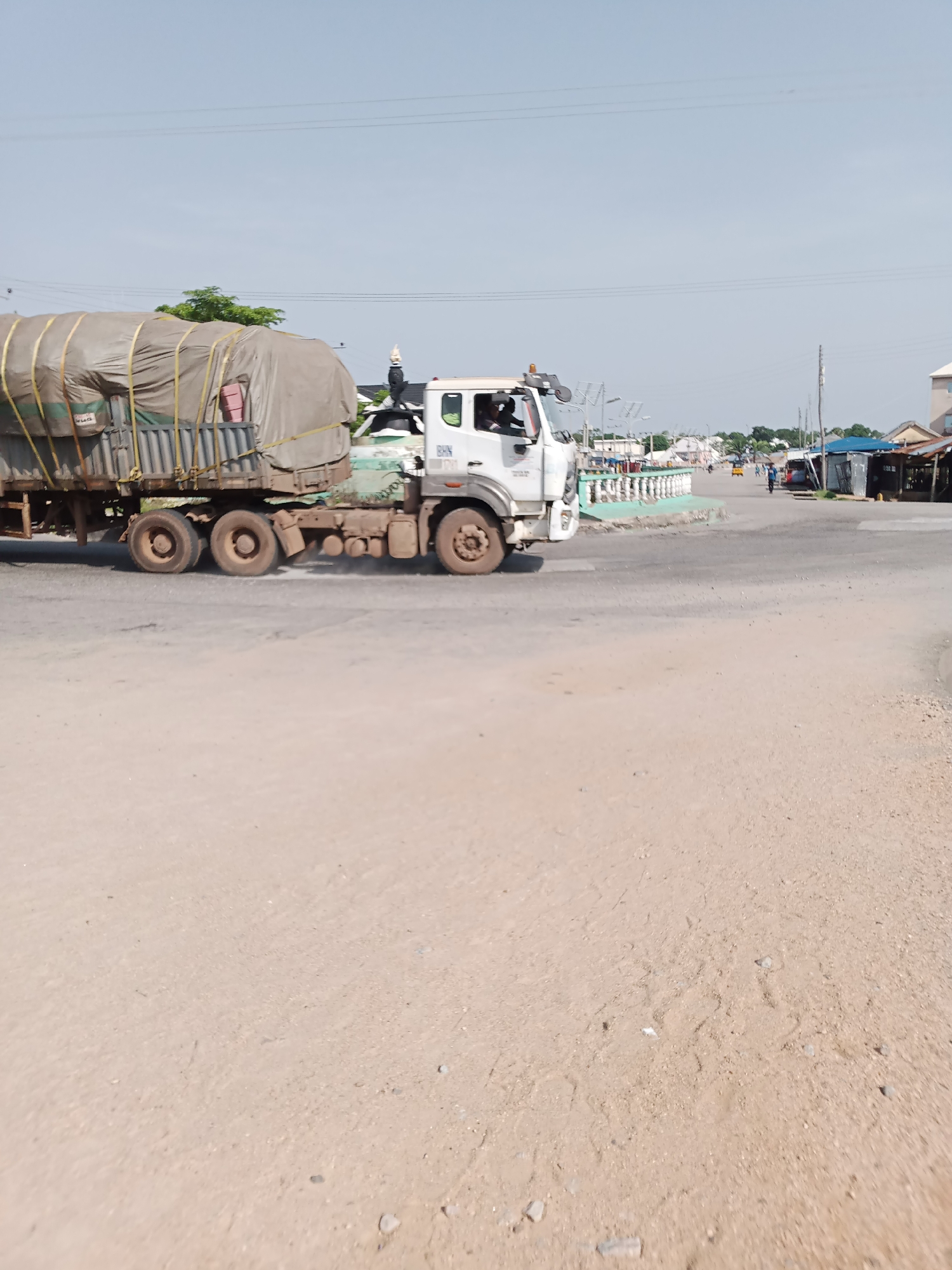 In Jalingo, Nigeria, a white truck carrying a large, tarp-covered load proceeds along a dirt road, its tires kicking up dust. The truck is loaded with what appears to be building materials, secured with yellow straps. The surrounding environment is a mix of arid land and scattered buildings, suggesting a developing or rural area. Several people are visible in the distance, some walking along the roadside and others near market stalls. A small building with a blue roof and zinc sheeting is visible on the right side of the road. The sky is clear and a bright blue.