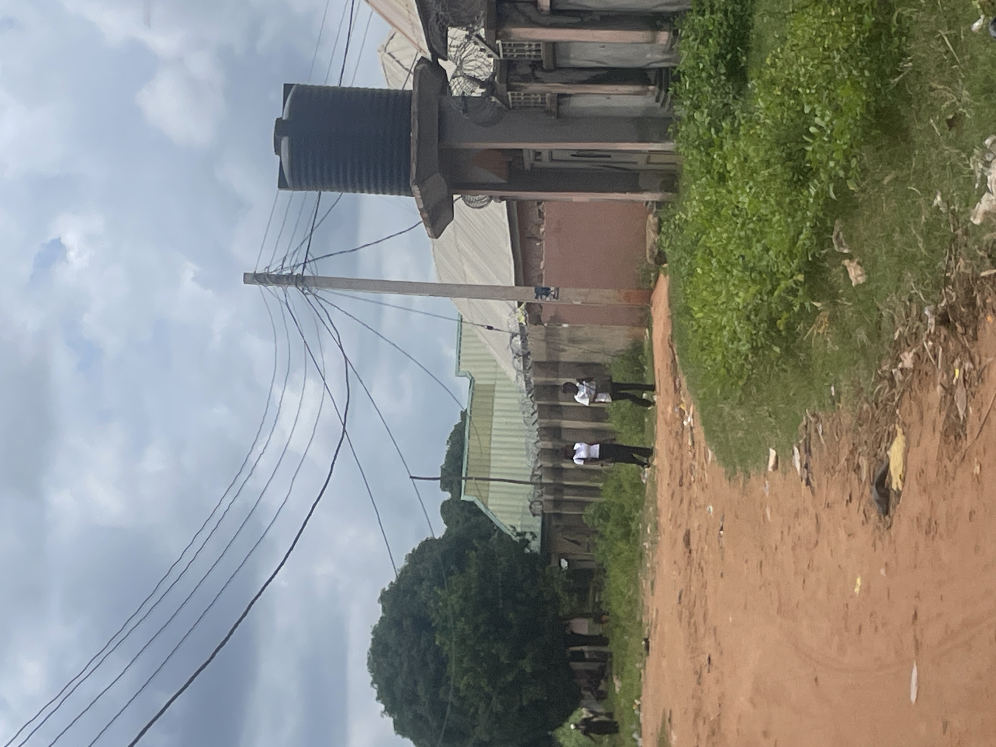 In Lafia, Nigeria, a dusty, unpaved road winds through a sparsely populated area under a cloudy sky. To the right, a large black water tank sits atop a concrete structure, suggesting a provision for water storage. Adjacent to it is a building with a corrugated metal roof and a fence topped with barbed wire, indicating security measures. Several individuals are present on the road. Two people in the foreground, one wearing a white top and black pants, and another in similar attire, walk away from the viewer. Further in the distance, on the left side of the road, another individual in dark clothing walks towards the left. A large, mature tree with dense foliage stands prominently on the left, near a cluster of buildings with metal roofs and fences. The environment appears to be a mix of residential and commercial structures, with evidence of limited infrastructure. The presence of litter scattered along the roadside indicates a lack of widespread waste management. The overall scene depicts a typical streetscape in a developing urban or peri-urban area, characterized by its simple construction, unpaved roads, and visible utility infrastructure.