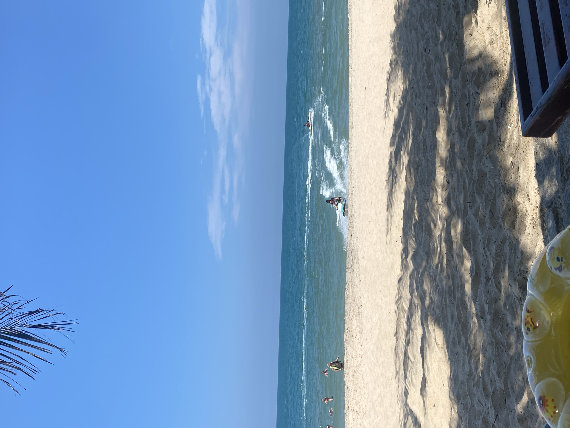 The image depicts a sunny day at a beach, likely in Phu Thuy Ward, Vietnam, as indicated by the clear blue sky and the presence of people enjoying water activities. The foreground is dominated by a sandy beach, with shadows of palm fronds cast across it, suggesting lush vegetation nearby. In the middle ground, the turquoise ocean meets the shore with gentle waves. Several individuals are visible in the water; some are wading or swimming, while others are riding jet skis, creating white wakes. One jet ski is in the process of turning, kicking up spray. A few people are standing on the sand at the water's edge. In the upper left corner, the tip of a palm frond hangs into the frame, framing the sky. The overall scene conveys a sense of leisure and recreation.