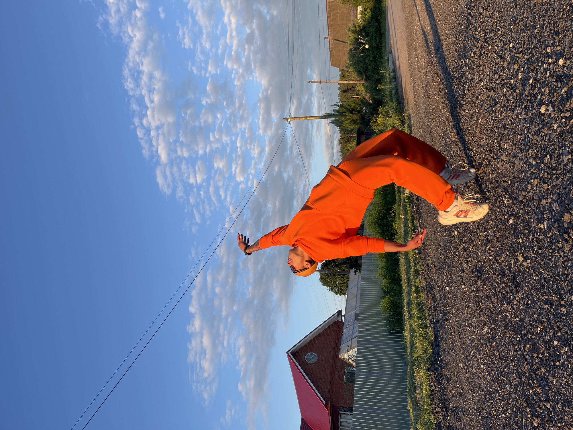 An individual is captured in a dynamic pose on a gravel road under a vast blue sky. The person, appearing to be an adult male, is dressed head-to-toe in a vibrant orange tracksuit, comprising a long-sleeved top and matching pants. They are also wearing white athletic shoes with orange accents and a pair of sunglasses or a hat. With arms outstretched and head tilted back, they are leaning backward, suggesting a moment of joy, freedom, or a playful stance.
The setting is an outdoor, possibly rural or semi-rural, landscape. A light-colored gravel road dominates the foreground, with patches of green grass and vegetation lining its edges. Overhead, multiple power lines crisscross the sky, supported by utility poles visible in the mid-ground. The sky is bright blue with scattered, fluffy white clouds that show hints of warm, golden light. This lighting, along with the long shadows cast on the road, indicates either late afternoon or early morning, commonly known as golden hour.
In the background, on the left side of the frame, a building with a red roof and a corrugated metal fence are visible. Another structure can be faintly seen further in the distance. The overall atmosphere conveyed is one of openness and natural light. No discernible text is visible within the image. The location context is Chaikovsky, Russia.