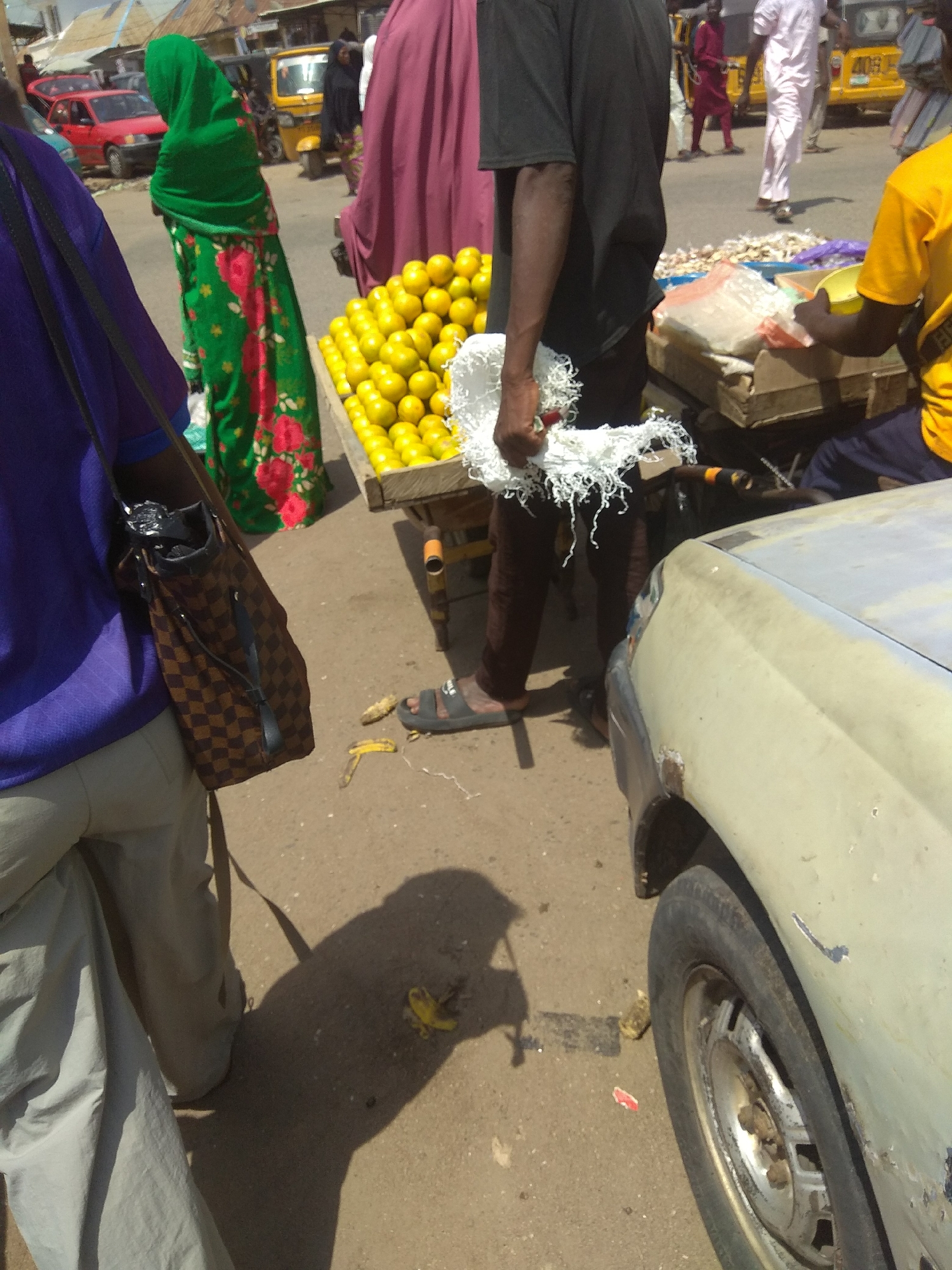 The image depicts a street scene in Jalingo, Nigeria, likely a market area, with several individuals present. In the foreground, a person wearing a purple shirt and light-colored pants is partially visible, carrying a brown and tan checkered bag. To their right, a person in dark clothing stands near a table laden with yellow fruits, possibly lemons or oranges. A white bundle of shredded material is held by the person in dark clothing, and another person wearing a yellow t-shirt is seated nearby.
Behind these individuals, a woman in a green hijab and a patterned dress walks away from the camera. Further in the background, a person in a maroon hijab is visible, and other figures can be seen near vehicles. A yellow auto-rickshaw is parked in the middle ground. The ground appears to be paved with asphalt, with scattered debris including banana peels and what looks like a crumpled red object. A light-colored car is parked in the right foreground, its front right wheel and fender visible. The overall impression is a busy outdoor market environment.