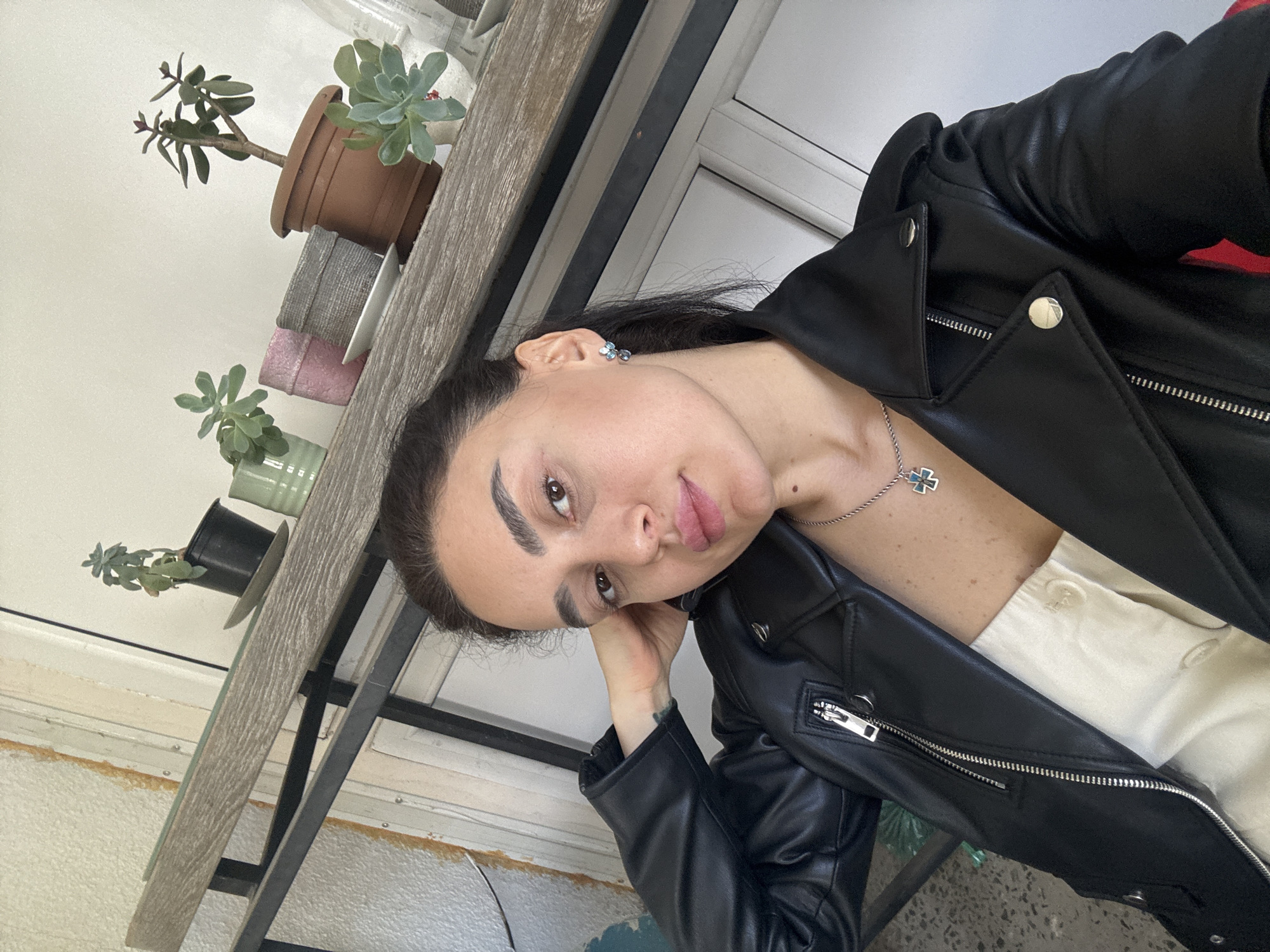 A woman with dark hair is posing indoors next to a window. She is wearing a black leather jacket over a white button-down shirt and a silver necklace with a blue cross. She has earrings and her hand is near her face. Beside the window are several small potted succulents on a gray shelf. The location is likely Tbilisi, Georgia, but this is not discernable from the visual information provided.