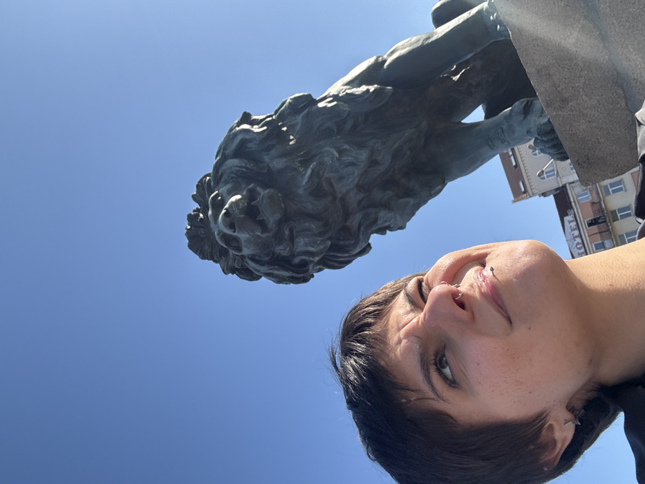 A low-angle outdoor photograph depicts a female individual alongside a bronze lion statue under a clear blue sky. The individual, positioned in the lower left, has short dark hair, a septum piercing, and a labret piercing, and is looking upwards and to the right with a playful expression, characterized by a scrunched nose and furrowed eyebrows. The large bronze lion statue, detailed with an open mouth suggesting a roar and a full mane, occupies the right side of the frame, situated on a stone plinth. In the background, a light-colored multi-story building with a partially visible "HOTEL" sign is discernible. The environment is illuminated by bright sunlight, casting direct light on the subjects. This scene is located at the Lion's Bridge (Lavov Most) in Sofia, Bulgaria.