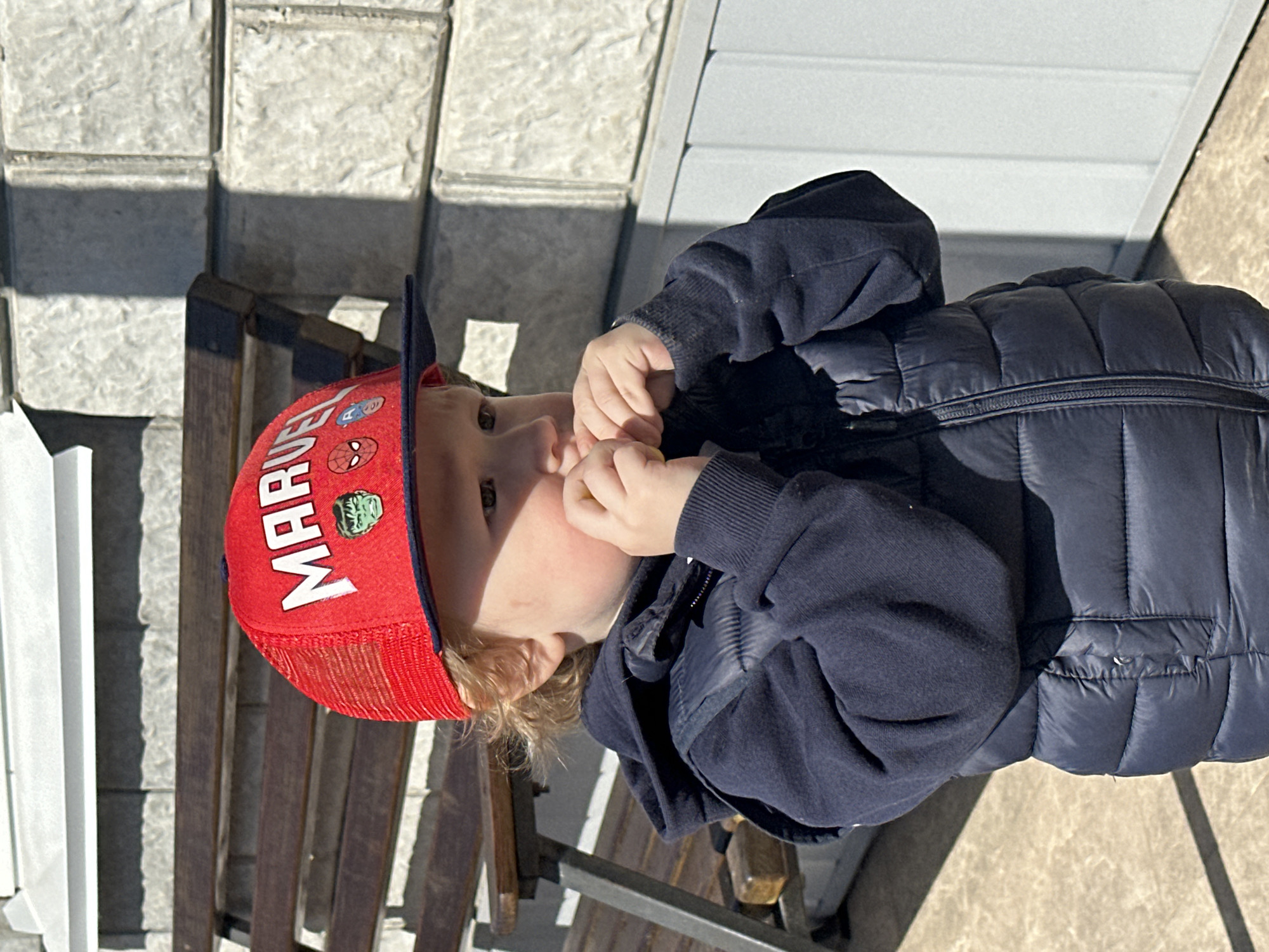 A child wearing a Marvel-themed red baseball cap, a dark blue hooded sweatshirt, and a dark blue puffy vest is seated on a park bench. The bench appears to be made of wood and metal, with dark vertical slats. The setting appears to be outdoors, possibly in Русский Вожой, Russia, given the prompt context. The child is looking to the left, and both hands are near his mouth. The environment includes a textured wall, likely made of stone blocks, and a light-colored siding of a building or structure is visible. There are shadows suggesting a sunny day.