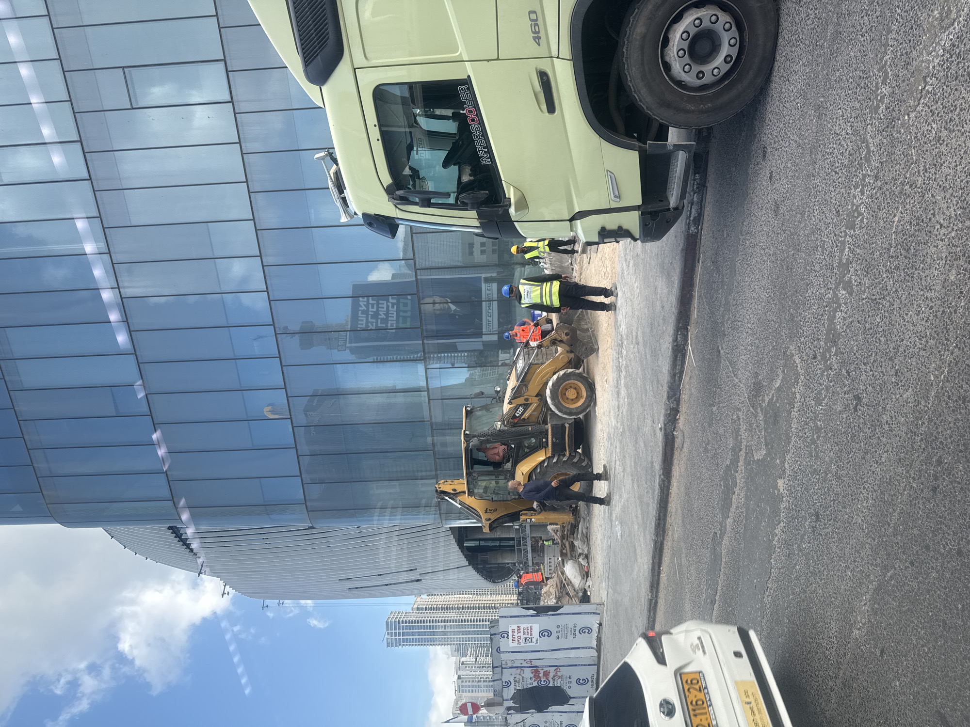 The image captures a construction scene in Givatayim, Israel. A large, pale yellow Volvo truck (model 460) is positioned on the right side of the frame, partially obscuring the view. In the center, a Caterpillar backhoe loader (model 432F) is parked amidst rubble, with construction workers nearby. One worker is inspecting the backhoe. Additional workers wearing reflective vests and hard hats stand near the truck, seemingly coordinating the activity. A building with a reflective glass facade rises prominently on the left, mirroring the sky and cityscape. The lower portion of the image reveals a white car with a license plate ending in "2-116-26" parked along the street. The background features a cluster of high-rise buildings in the distance, indicative of Givatayim's urban landscape. Signage with Hebrew text is visible near the white vehicle, cautioning about the construction site.