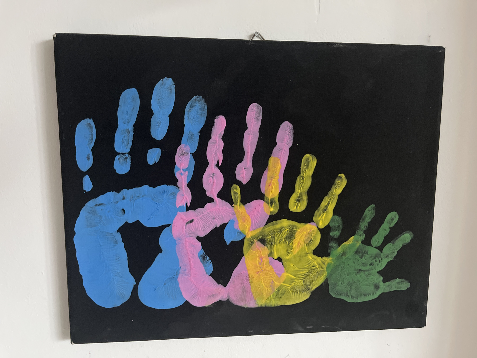 A framed canvas artwork depicting multicolored handprints against a black background is mounted on a white wall. The handprints are in blue, pink, yellow, and green, arranged in overlapping layers. The canvas is hung with a silver hook. The scene appears to be indoors, likely in a residential setting, with the white wall indicating a typical interior finish. The overall impression is that of a piece of children's art or a family keepsake. There is no indication of any specific location within Kohtla-Järve linn, Estonia, as the image contains no discernible landmarks or identifying features of that city.