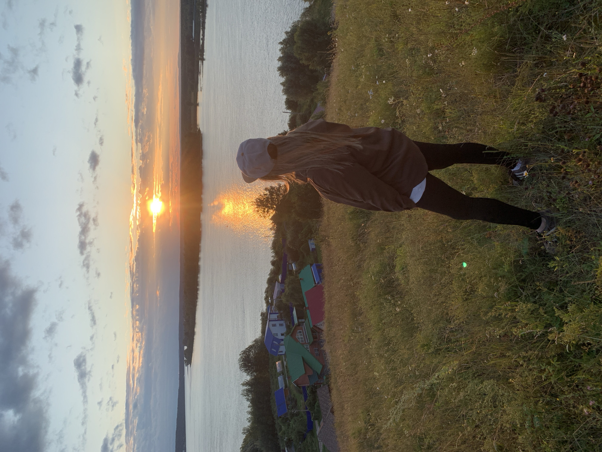 A person stands on a grassy hill, facing a wide river at sunset. The person is wearing a brown hooded sweatshirt, black leggings, a gray baseball cap, and sneakers. The sun is setting over the river, casting a golden glow on the water and clouds. In the background, there is a small village with houses and trees along the riverbank. The grassy hill is covered in dry grass and wildflowers. The sky is a mix of blue and orange, with scattered clouds. The location is the city of Olkhovka, Russia.