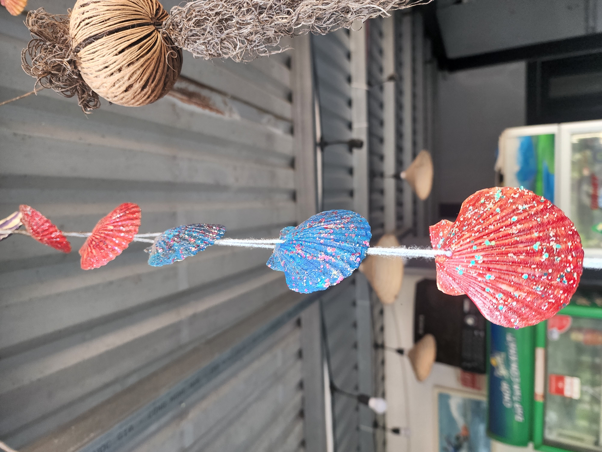 This image captures a close-up perspective of decorative items hanging from a ceiling in an indoor or covered outdoor space in Phan Thiet Ward, Vietnam. In the immediate foreground, a vertical string displays five painted seashells, alternately colored red/orange and blue, each adorned with glitter. The largest shell, at the bottom, is red/orange with numerous blue and white glitter specks, and is in sharp focus. Progressing upwards, two blue shells and two more red/orange shells are visible, with clarity decreasing towards the top. To the right, a second hanging decoration consists of a spherical, light-brown object, likely a dried seed pod or woven natural fibers, from which a substantial mass of dried, stringy, light-brown plant material hangs. The background, rendered with significant blur, features a light-grey corrugated metal ceiling with parallel horizontal ridges. Below the ceiling, multiple pendant lights are discernible, some with bare bulbs and others with conical, light-colored shades. In the lower-left background, a green commercial display cooler or refrigerator is partially visible, topped by a framed picture depicting a blue and white landscape scene.