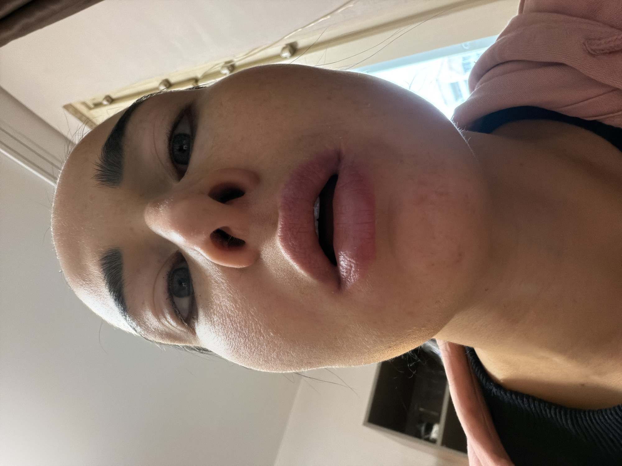 A close-up, upward-facing shot of a person's face. The person has fair skin, dark eyebrows, and dark eyes. Their lips are parted slightly. The background is blurred, showing a window with natural light coming through, and parts of a room including what appears to be part of a cabinet. The person is wearing a black top and a pink hoodie. No information in the image allows for locating it in St. Petersburg, Russia.