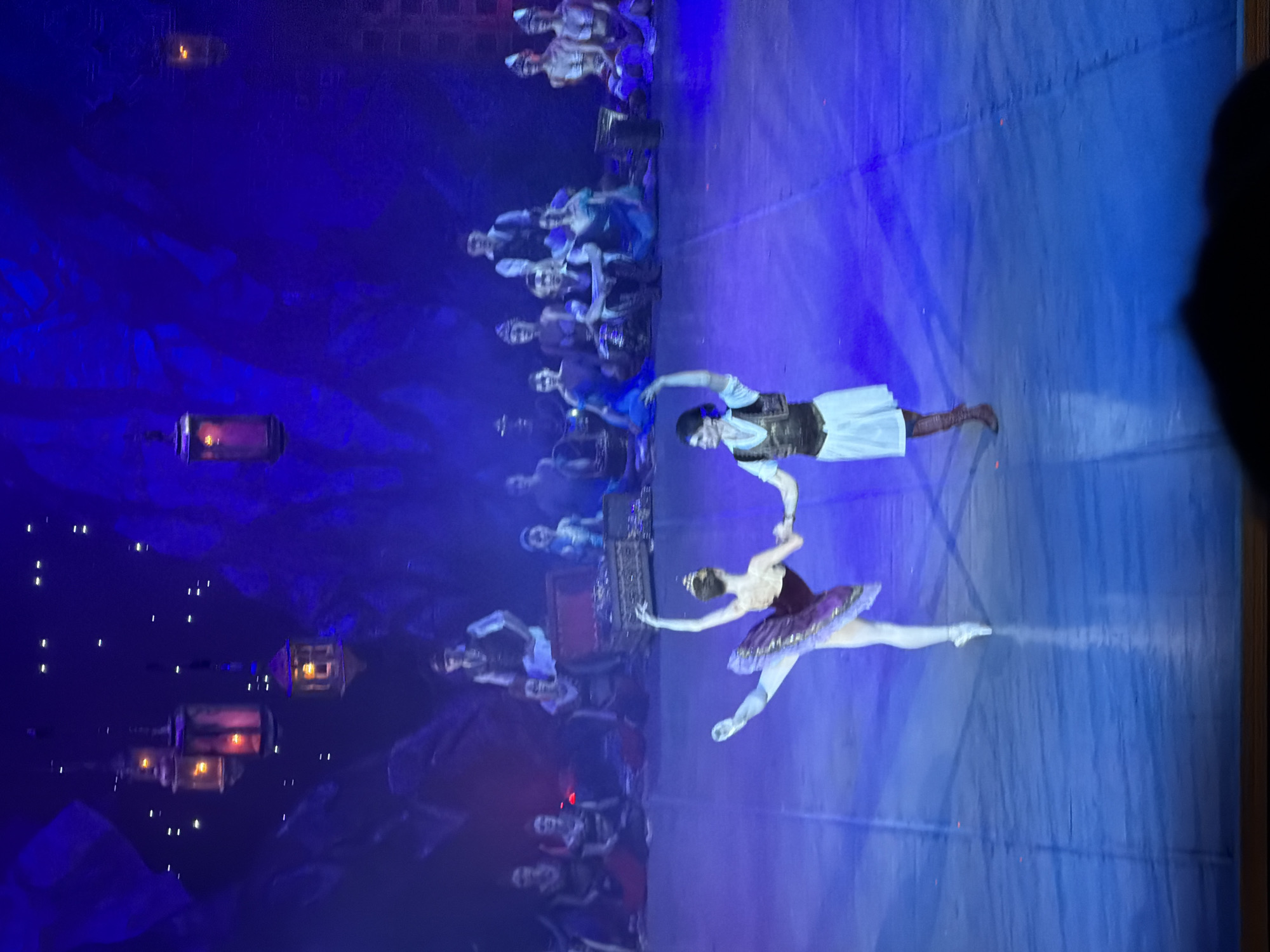 The image depicts a ballet performance in what appears to be the city of Izhevsk, Russia. Two principal dancers, a male and a female, are performing center stage under blue lighting. The female dancer wears a dark purple tutu and is en pointe, while the male dancer is in costume with a vest and white sleeves. They are holding hands, performing a lift. A chorus line of dancers and audience members sit behind them, and there are theatrical elements, including painted backdrops, and lanterns as part of the stage production.