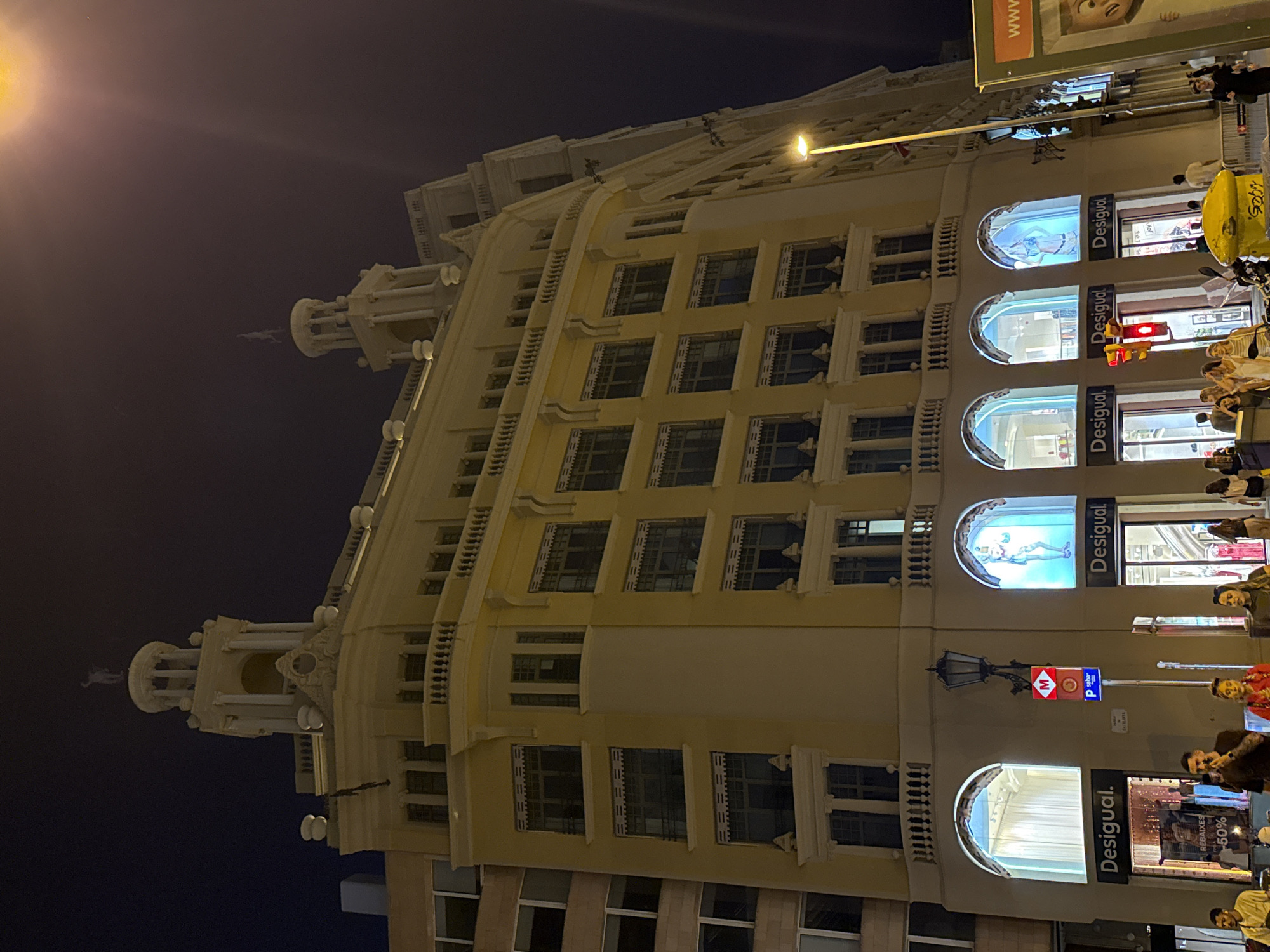 The image shows the facade of a multi-story building at night in Barcelona, Spain. The building is predominantly yellow and features ornate architectural details, including balconies, arched windows, and decorative elements on the roofline. The ground floor has multiple storefronts with the "Desigual" logo prominently displayed. Some of the shop windows feature illuminated displays of mannequins. Several people are walking on the sidewalk in front of the shops. Traffic lights and street lamps are visible, indicating an urban environment. A yellow street lamp with a bright light is also prominent on the right side of the image. The sky is dark, suggesting it is nighttime.