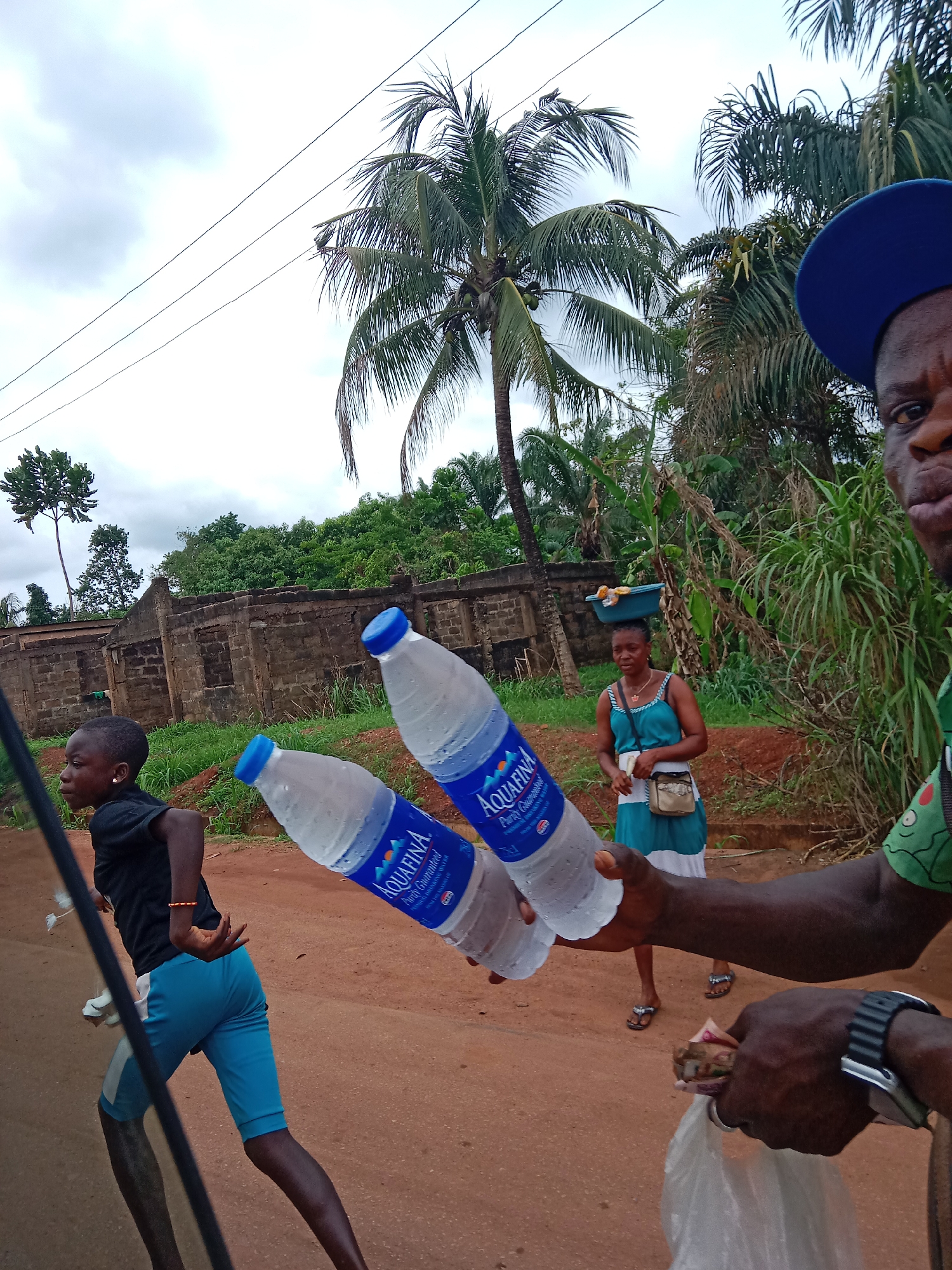 The image, taken in Oji River, Nigeria, shows a man holding two bottles of Aquafina water, a young child running on the left, and a woman carrying a container on her head in the background. The road is a red-brown color, indicating a laterite-rich soil common to the area. The surrounding environment includes palm trees, dense foliage, and a low brick wall. The weather appears overcast.