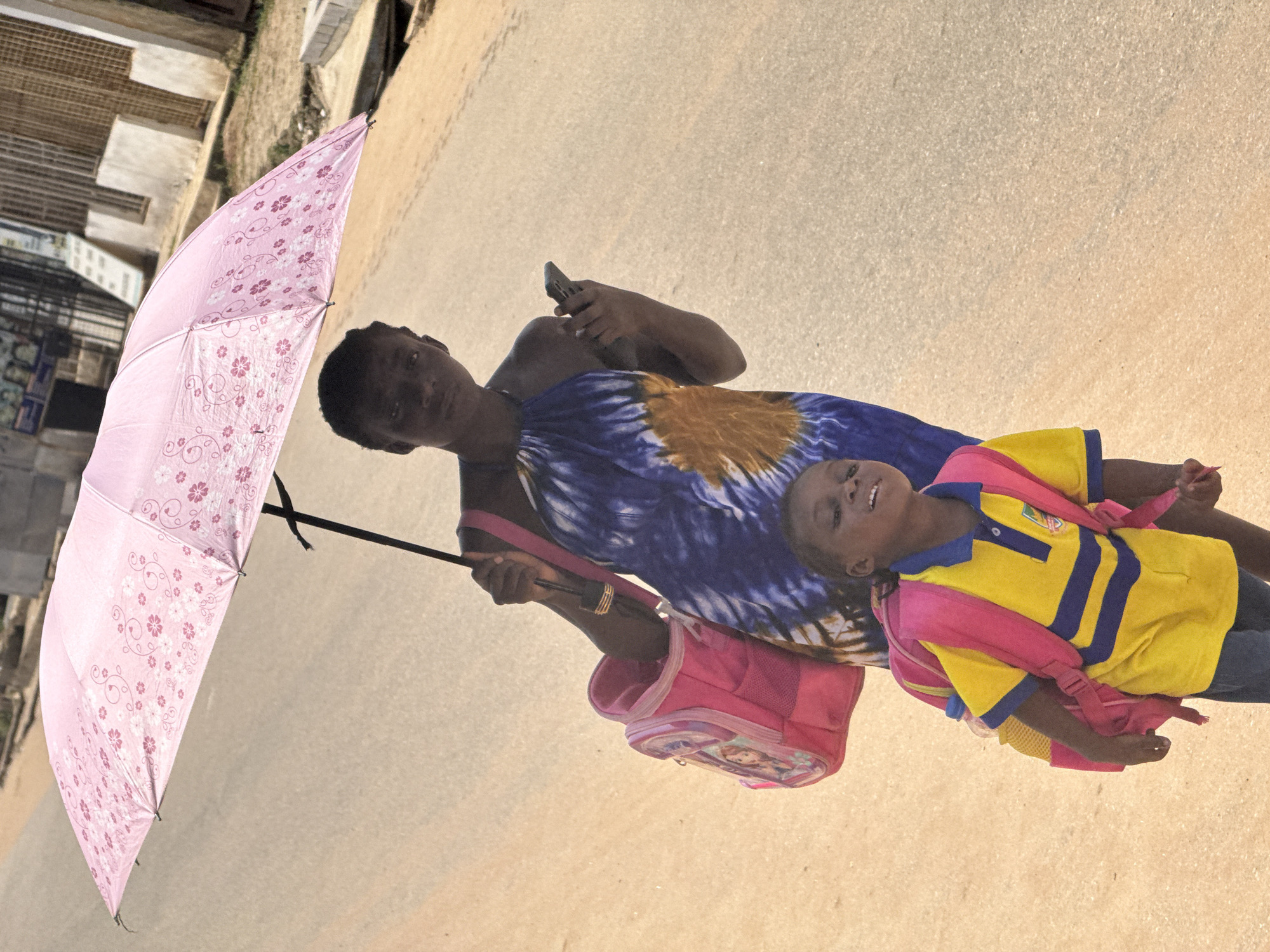 A young Black child wearing a yellow polo shirt with blue stripes and a pink backpack is walking on a paved street in Abuja, Nigeria. The child is looking up and to the left with a smile. Behind the child, a Black woman wearing a blue and orange tie-dye dress and holding a pink umbrella and a cellphone is walking. The woman is holding the umbrella over both herself and the child. A pink backpack is also hanging from the woman's left shoulder. The environment appears to be a street in a populated area, with buildings visible in the background. The lighting suggests it is daytime, with strong sunlight casting shadows.