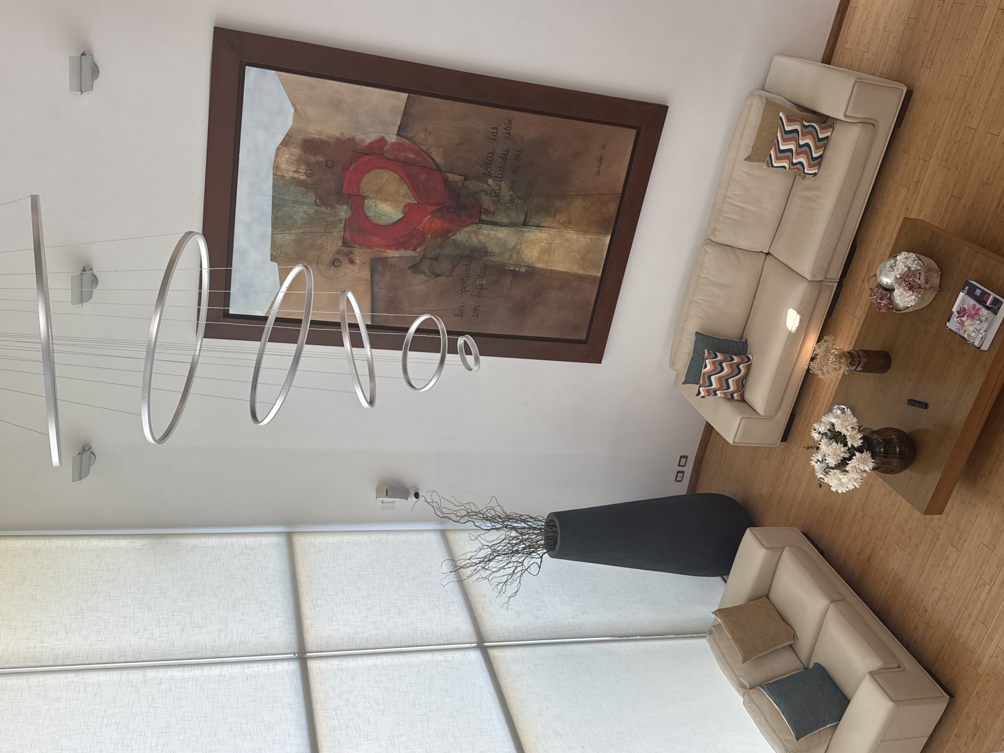 The image depicts a modern living room in San Bernardino Tlaxcalancingo, Mexico, viewed from an elevated perspective. The room features two light beige sofas, one elongated and another smaller, arranged around a wooden coffee table. The table is adorned with flowers, books, and vases. A large abstract painting with a dark brown frame hangs on the wall, adjacent to a contemporary chandelier with circular elements. Below the chandelier, a floor-to-ceiling window with white blinds provides natural light. A tall, dark-colored vase filled with branches stands in front of the window. The floor is wooden.