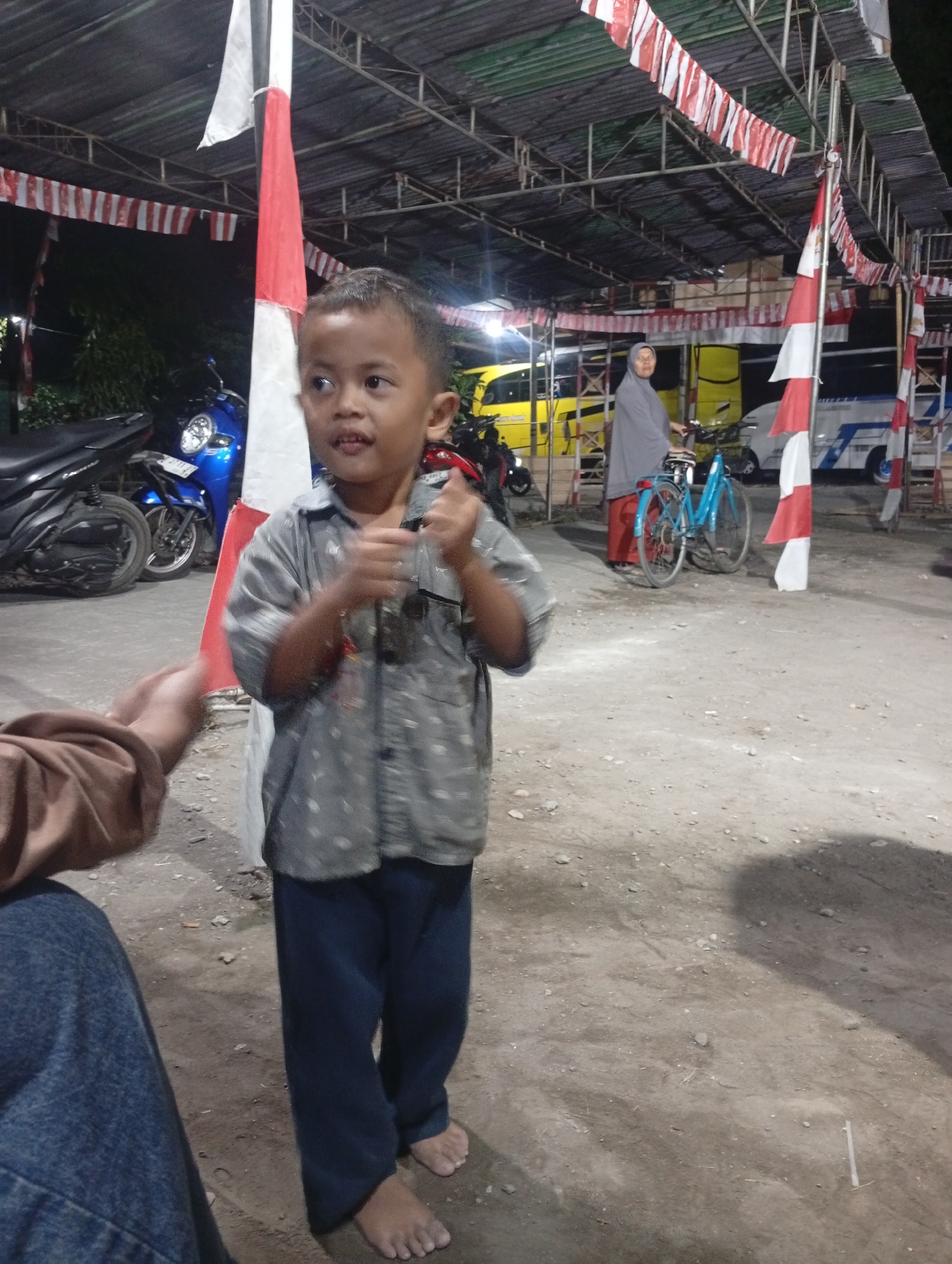 In Sewon, Indonesia, a young boy stands barefoot on a dirt ground, wearing a grey patterned shirt and dark blue pants. He is looking to his right, his hands slightly raised in front of him. To his left, another person's arm and shoulder are visible, wearing a brown shirt and blue jeans. Behind the boy, a woman in a grey hijab and red sarong is sitting on a blue bicycle. In the background, several parked motorcycles and vehicles are visible, including a yellow bus and a white bus. Red and white striped banners are hung overhead, and a large red and white flag is prominently displayed on a pole. The scene appears to be outdoors under a covered structure, likely a market or event space, with artificial lighting contributing to the evening atmosphere.
