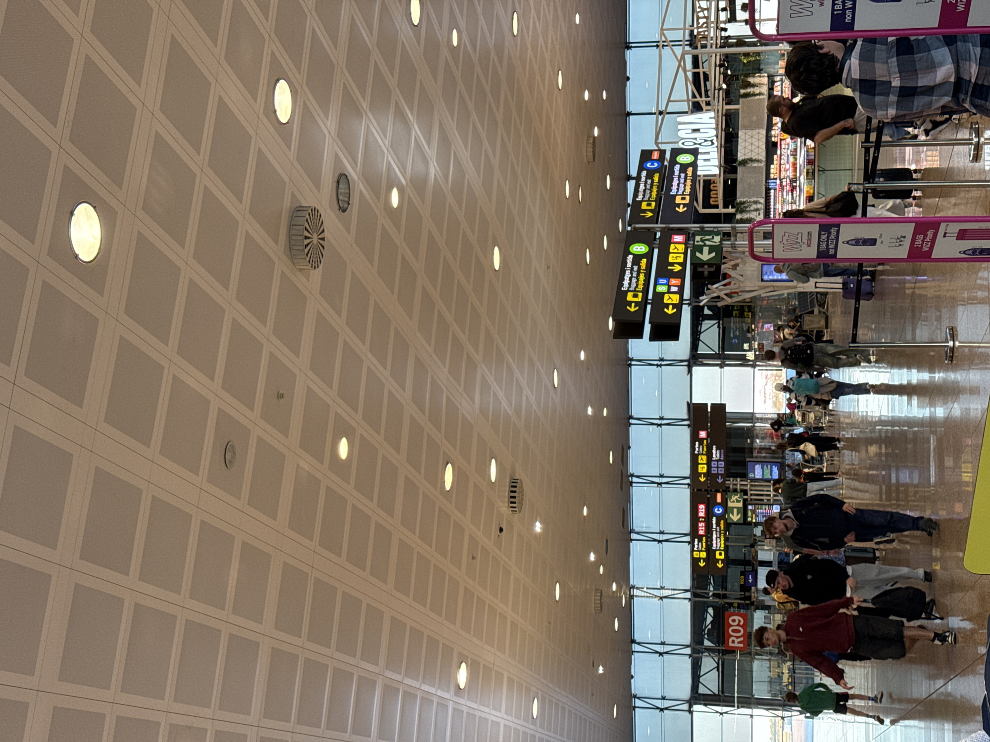 The image shows the interior of an airport terminal, likely Barcelona–El Prat Airport in El Prat de Llobregat, Spain. The perspective is skewed, with a focus on the ceiling and the right side of the image. The ceiling is composed of square panels with embedded circular lights and some ventilation grilles. The terminal has tall glass walls allowing natural light into the space.
People are visible in the terminal. Some are standing and walking, while others are in line. There are directional signs hanging from the ceiling to guide passengers. Specifically, signs direct people to gates R09 and R15-R19, and the signs also include directions for baggage claim.
A Wizz Air check-in line is also visible with signage for “1 Bag Only non WIZZ Priority” and “2 Bags WIZZ Priority.”
