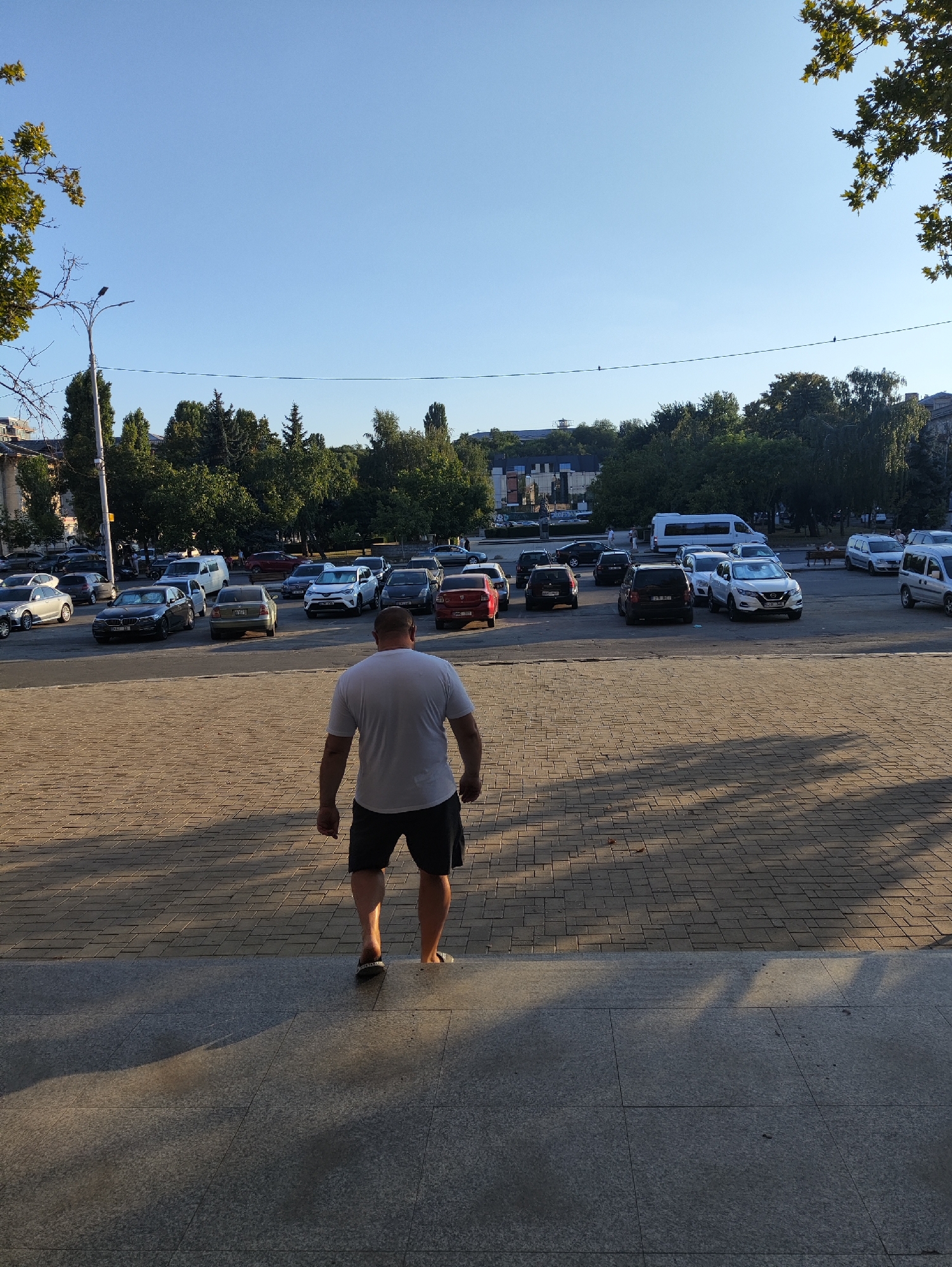 A man wearing a white t-shirt and black shorts walks down a paved stairway in a city square. He is facing away from the camera and towards a parking lot filled with various cars, including a white van, a white SUV, and several sedans. Trees line the background, with buildings visible behind them. The sky is clear and blue, indicating a sunny day. The lighting suggests the photograph was taken in the late afternoon or early evening, given the long shadows cast by the man and the trees. This scene is likely in the city of Chișinău, Moldova, given the context provided by the user's prompt.