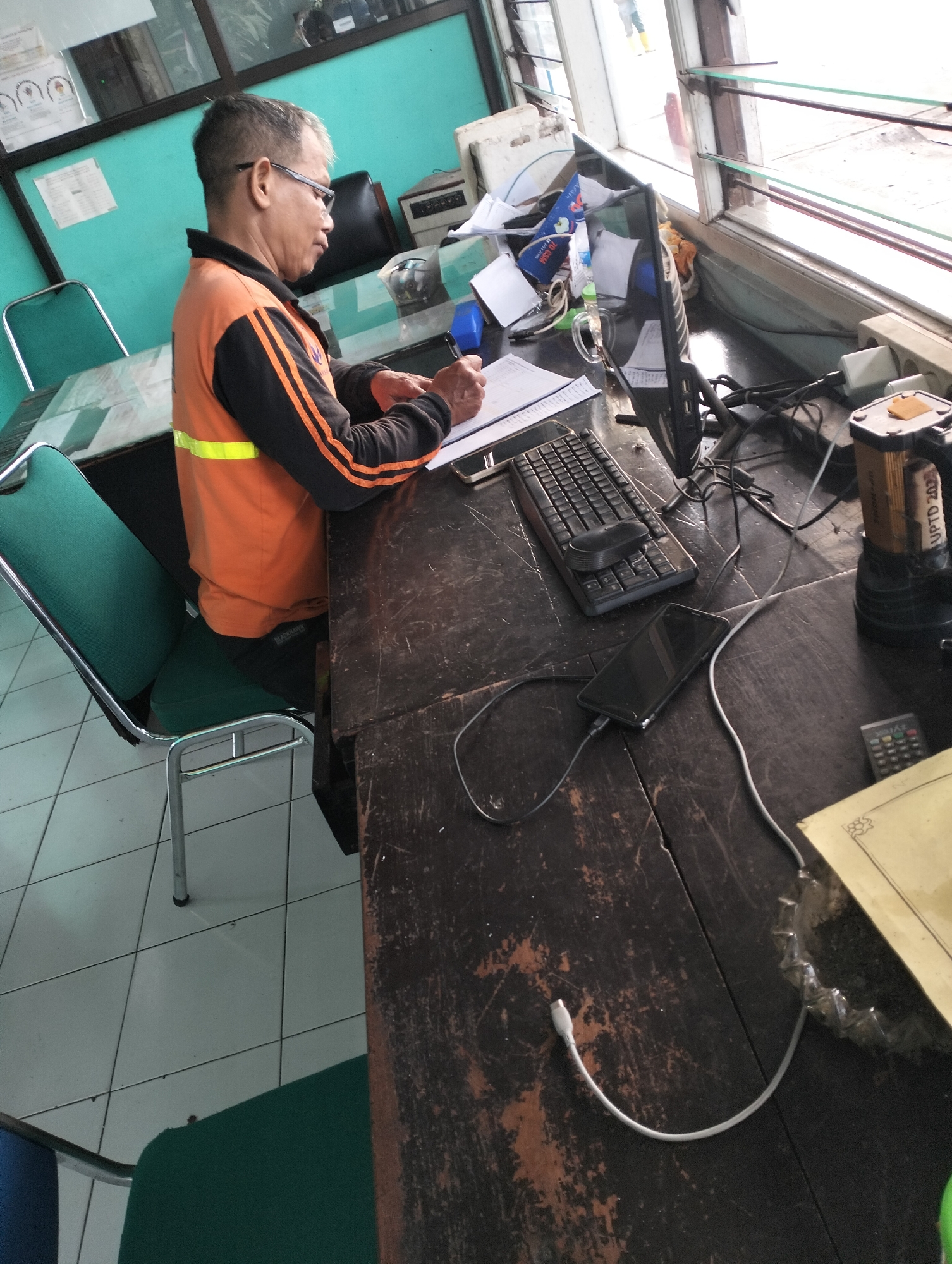 An adult Indonesian male wearing glasses and an orange and black long-sleeved shirt with a yellow reflective stripe is seated at a dark wooden desk, writing on a document. The desk is cluttered with various items, including a computer monitor, keyboard, mouse, a smartphone connected to a charger, a flashlight, and a remote control. The man is positioned in what appears to be an indoor workspace, possibly a security post or an administrative office, with a window overlooking an outdoor area. In the background, there are teal-colored walls, a filing cabinet, and two green chairs. The floor is tiled in a white and light gray checkered pattern. The scene suggests a work environment in Surakarta, Indonesia, where the man is engaged in administrative tasks.