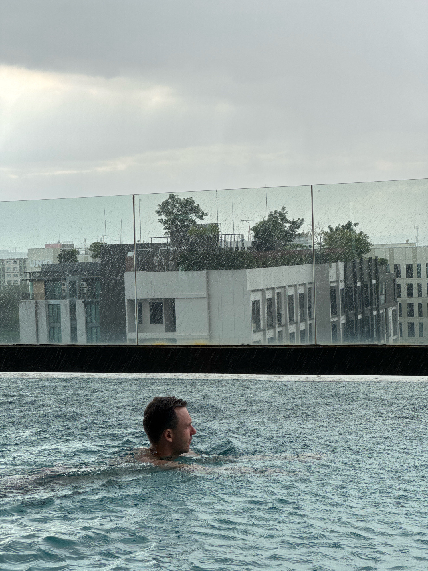 In the Samrong Nuea Subdistrict Municipality of Thailand, under a cloudy, overcast sky with visible rain, a man is swimming in an infinity pool. The water surface is disturbed by raindrops, creating ripples. The man is positioned in the foreground, facing to the right, with his head and shoulders above the water. Behind him, a glass railing separates the pool from a cityscape. The city skyline features buildings of varying architectural styles and heights, some with visible windows and balconies. Trees are also present on some of the building rooftops. The overall scene suggests an urban environment experiencing inclement weather.