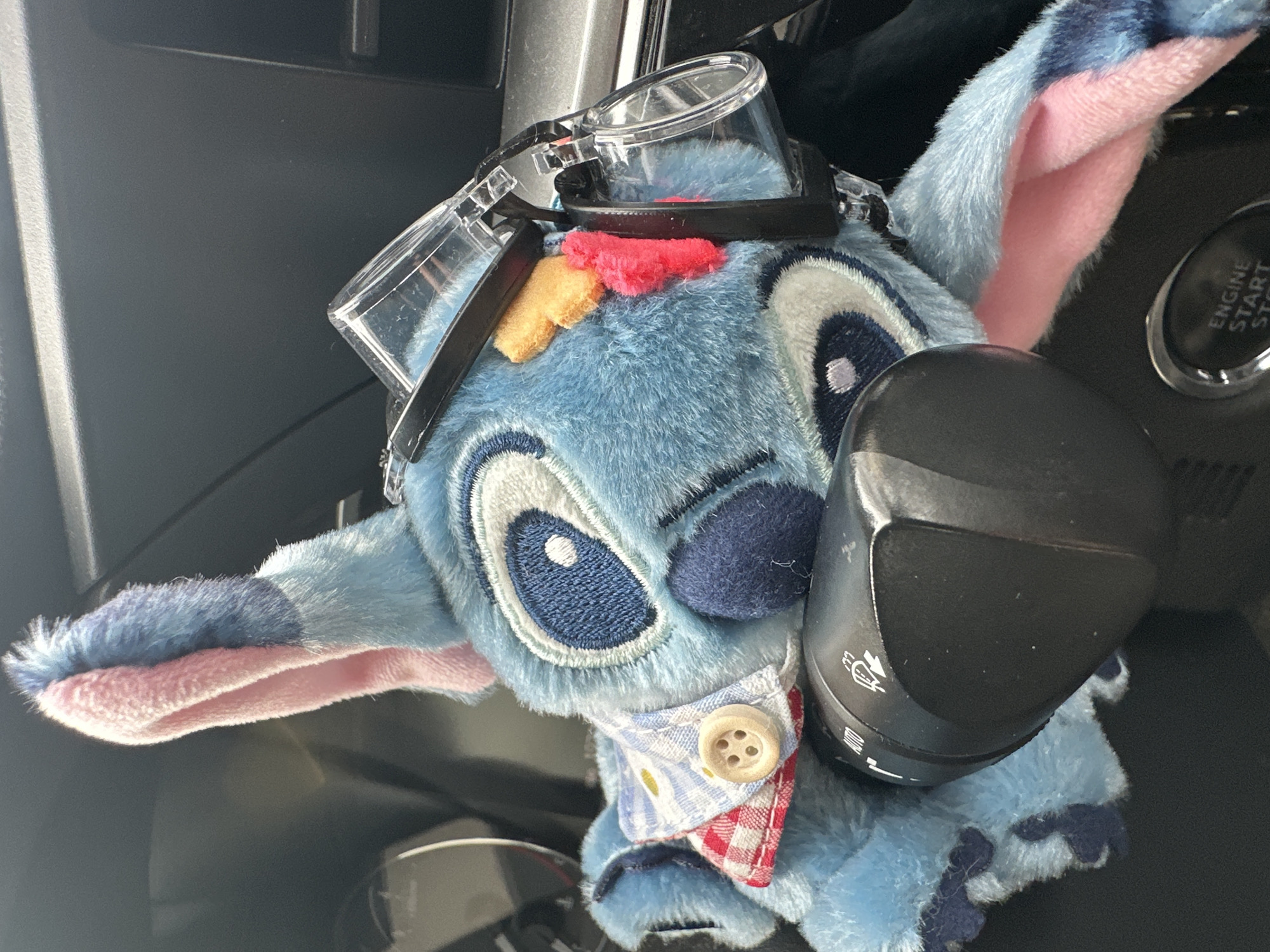 A plush toy of Stitch from Lilo & Stitch is in the interior of a car. Stitch has goggles on his head and a button-down shirt. Stitch is in front of a turn signal lever and an engine start button. The location is unknown and therefore the image cannot be said to be in Chaikovsky, Russia.