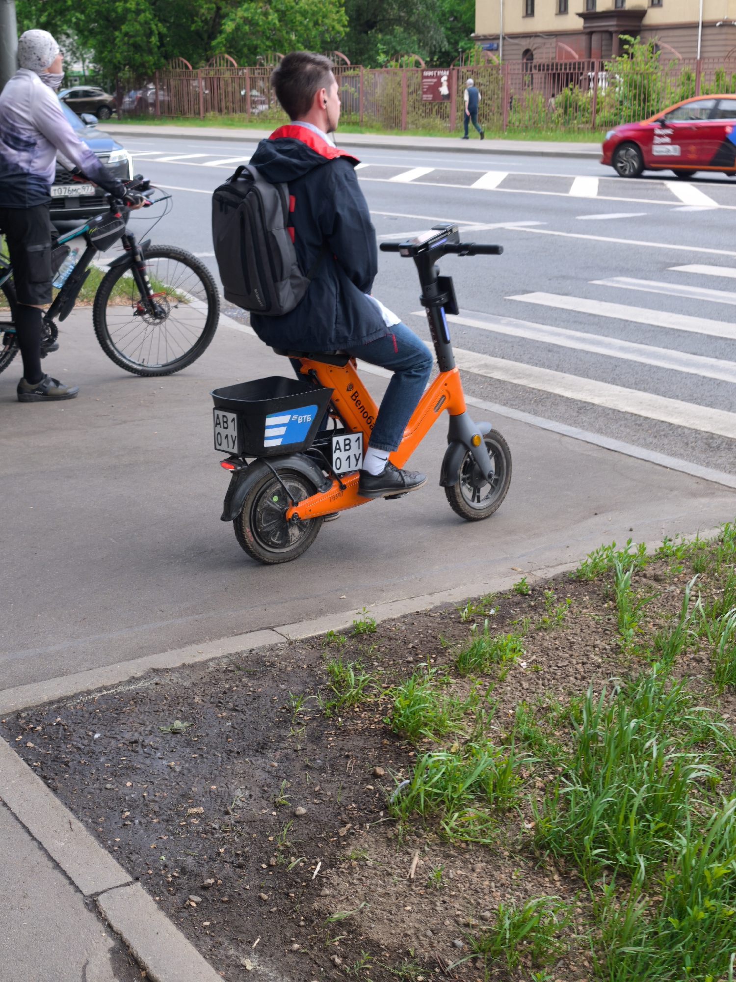 A daytime image captures a man riding an orange electric scooter on a sidewalk, likely in Moscow, Russia. He wears a dark blue jacket, jeans, and a gray backpack. The scooter has a black basket on the back with the license plate "AB1 01Y." The Cyrillic word "Велобайт" and a "ВТБ" logo are visible on the scooter. A cyclist in a light-colored jacket stands next to a bicycle on the same sidewalk. A crosswalk is visible behind the scooter, leading to a street where a red car and a person walking on the sidewalk can be seen. A building with a reddish-brown exterior and multiple windows is in the background. The sidewalk is adjacent to a patch of soil with grass growing on it.