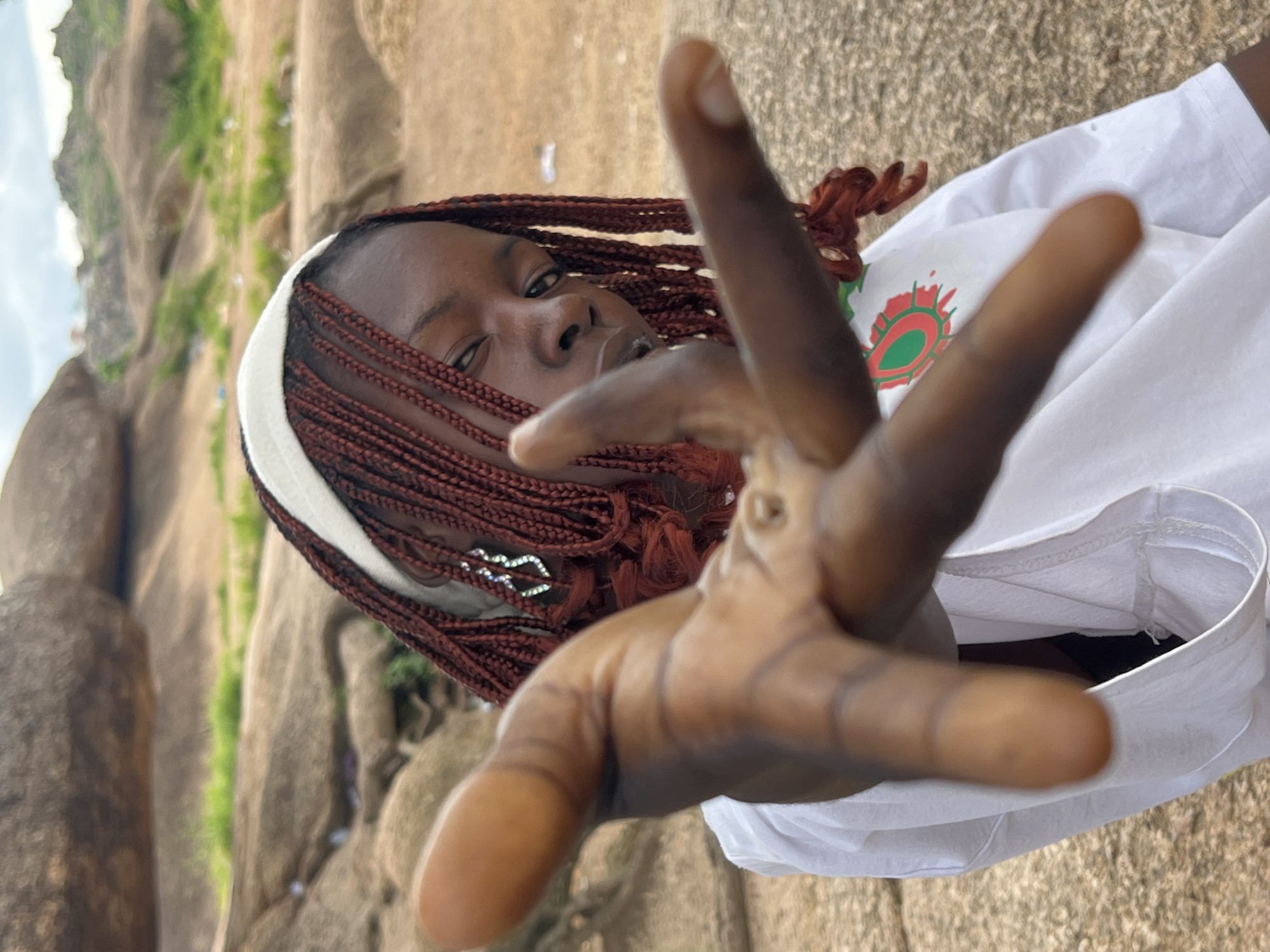 A young Black female with dark skin and braided auburn hair, wearing a white headband and a white t-shirt with a green and red circular logo, reaches her right hand forward toward the camera. She is positioned outdoors, in what appears to be a rocky, natural landscape. The background is dominated by large, weathered rock formations and sparse green vegetation. The sky above is overcast, suggesting a cloudy day. The location is identified as the Shere Hills in Jos, Nigeria, a region known for its granite rock formations and savanna environment. The image is captured in a medium close-up, with the woman's hand prominently in the foreground, slightly out of focus, obscuring part of her face.