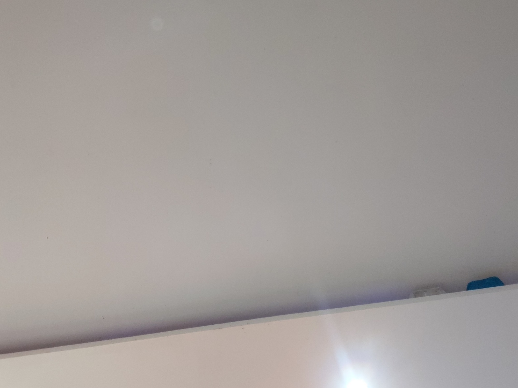 This image depicts an indoor scene, primarily focused on a light-colored, uniform surface, likely a ceiling or upper wall, which occupies the majority of the frame. The surface appears off-white or light gray with a smooth texture, showing only minor dust specks. A distinct horizontal line near the bottom separates this upper surface from a lower, brighter white surface, suggesting the top edge of a shelf, cabinet, or another wall. A faint purple hue is visible along this dividing line, potentially due to ambient lighting or reflection.
In the bottom right corner, resting on the edge of the lower white surface, are two small objects. One is a clear or translucent white rectangular item, and next to it is a small, rounded blue object. A bright light source, possibly artificial or a window with strong sunlight, creates a noticeable lens flare emanating from the bottom right corner. No people or specific activities are visible. The scene is static and appears clean and minimally furnished. There is no discernible text within the image. The location context for this indoor scene is Santa Ana, Venezuela.