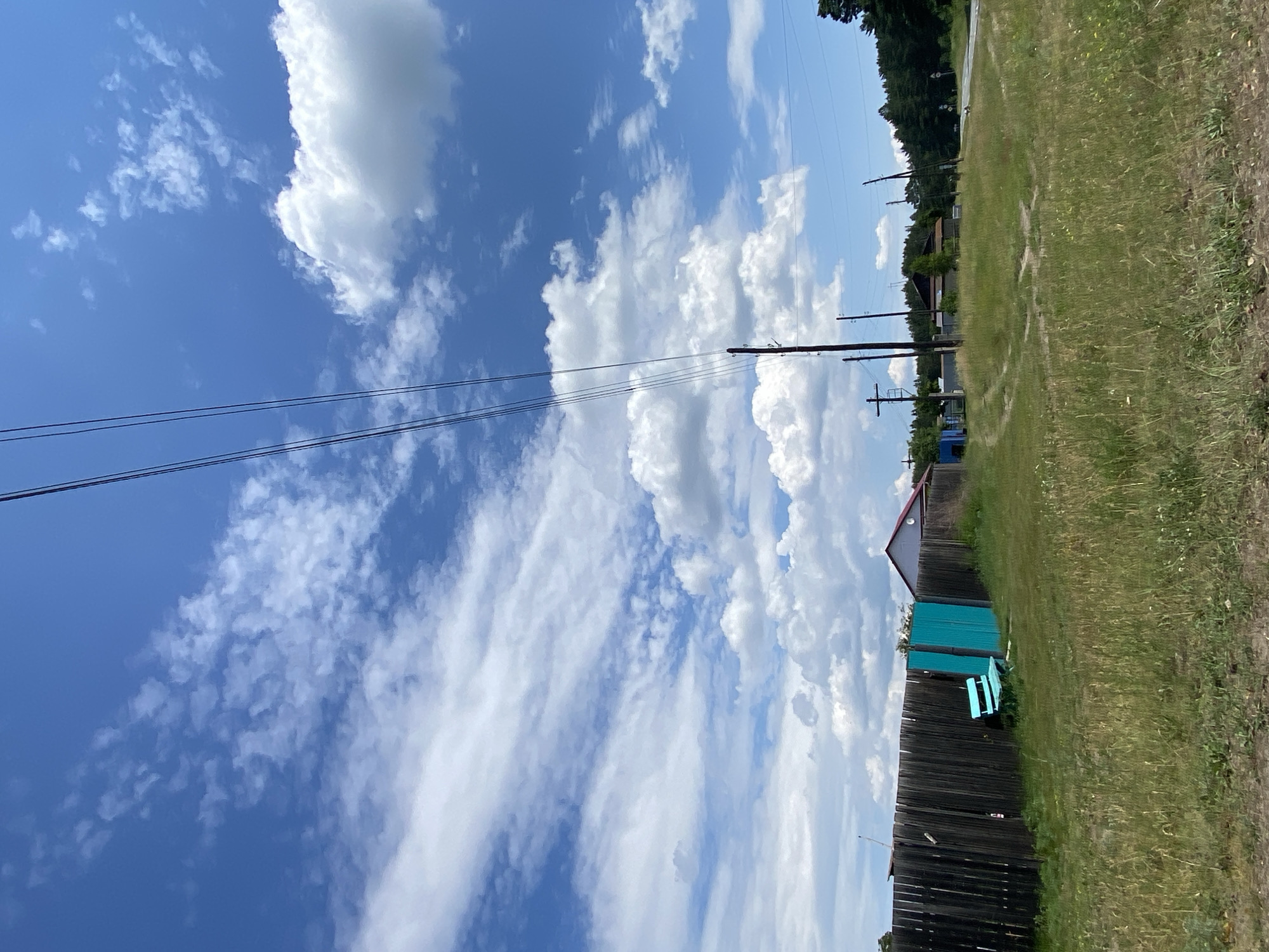 This is a medium-shot, eye-level, outdoor, daytime, vertical, full-color photograph taken in the city of Znamensk, Russia. The image depicts a rural scene with a wooden fence in the foreground, a bright teal-colored gate and bench within a grassy area. Beyond the fence, several power lines stretch diagonally across the bright blue sky, which is dotted with white cumulus clouds. A few wooden utility poles are visible, supporting the lines. Further in the background, there are residential buildings with red roofs and a dense forest line the right side of the frame. A dirt road with tire tracks curves through the grassy landscape. The overall impression is one of a quiet, rural setting on a sunny day.